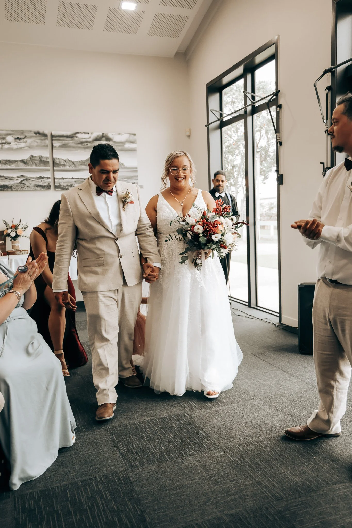 A bride and groom holding hands during their wedding ceremony inside a bright room with large windows. The bride is in a white wedding dress holding a bouquet and smiling, while the groom is dressed in a beige suit and bow tie with a serious expressi