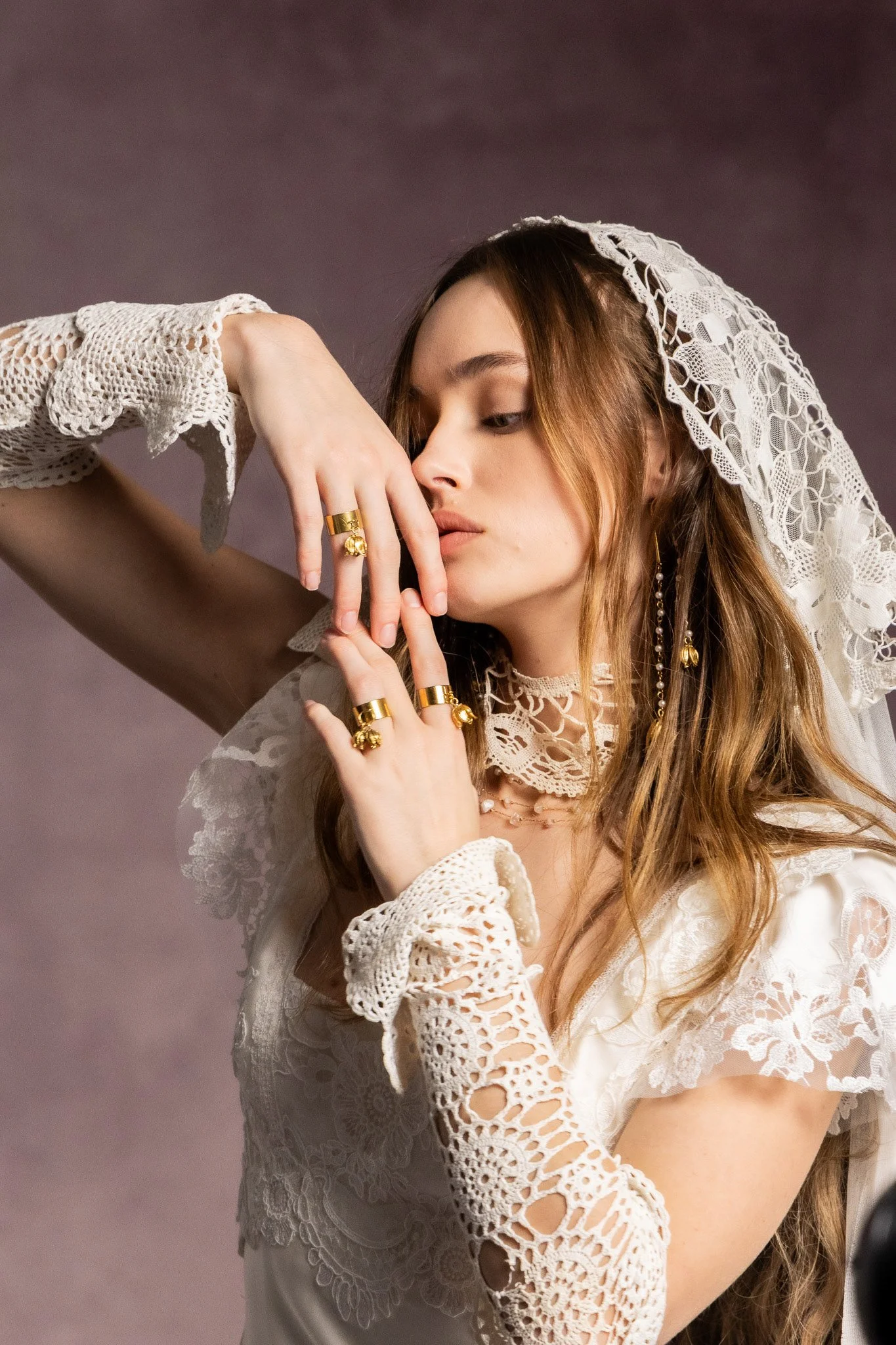 A woman with long, wavy, reddish-brown hair, wearing a white lace blouse with intricate cutouts, a lace headscarf, and multiple gold rings and earrings, is posing against a plain background, with her hand near her face.