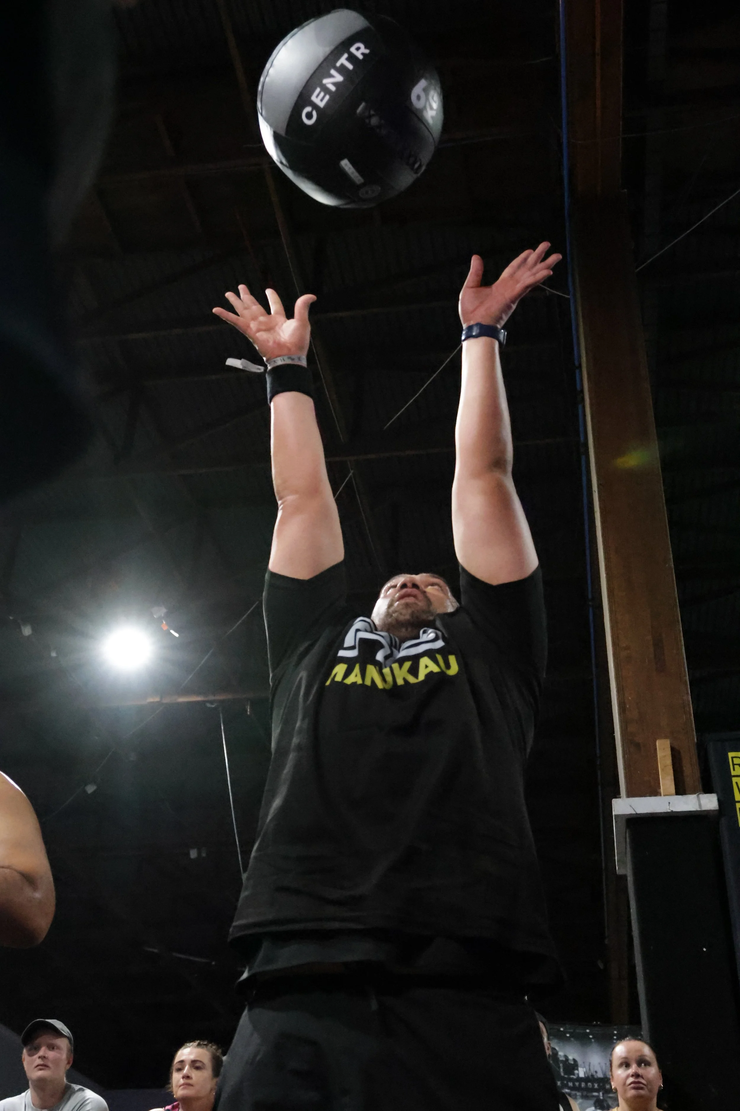 A man with a beard and black shirt reaching up with both hands towards a black and white medicine ball labeled 'CENTR'. Several people are in the background, watching in an indoor gym.