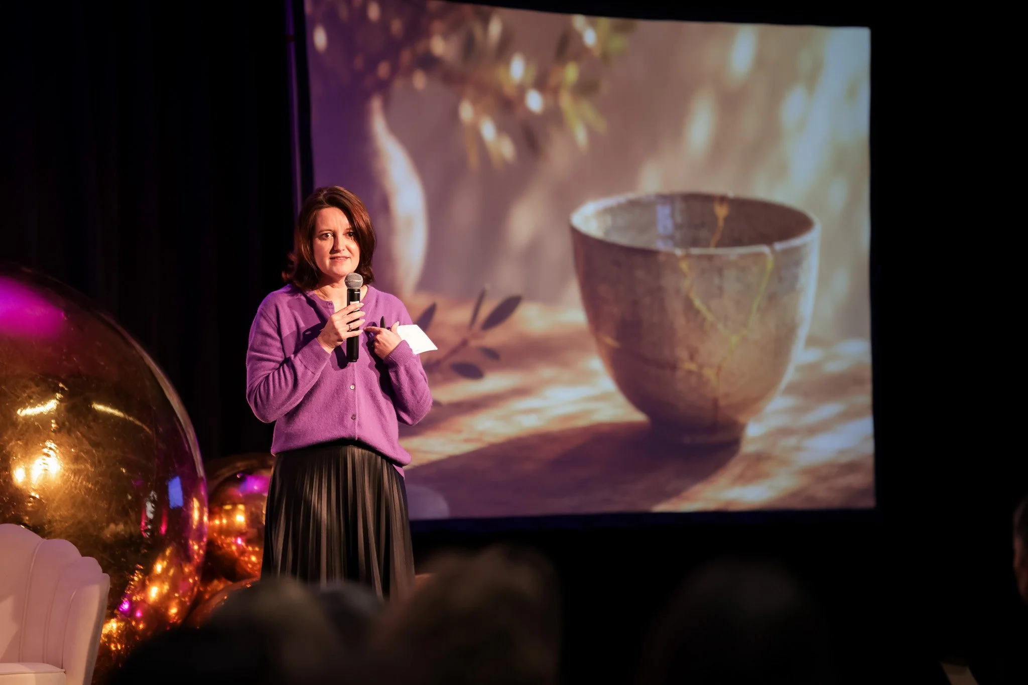A woman with shoulder-length brown hair, wearing a purple cardigan and striped skirt, holding a microphone and standing on stage during a presentation or event. Behind her, a large screen displays an image of a ceramic cup that is cracked, with a blu