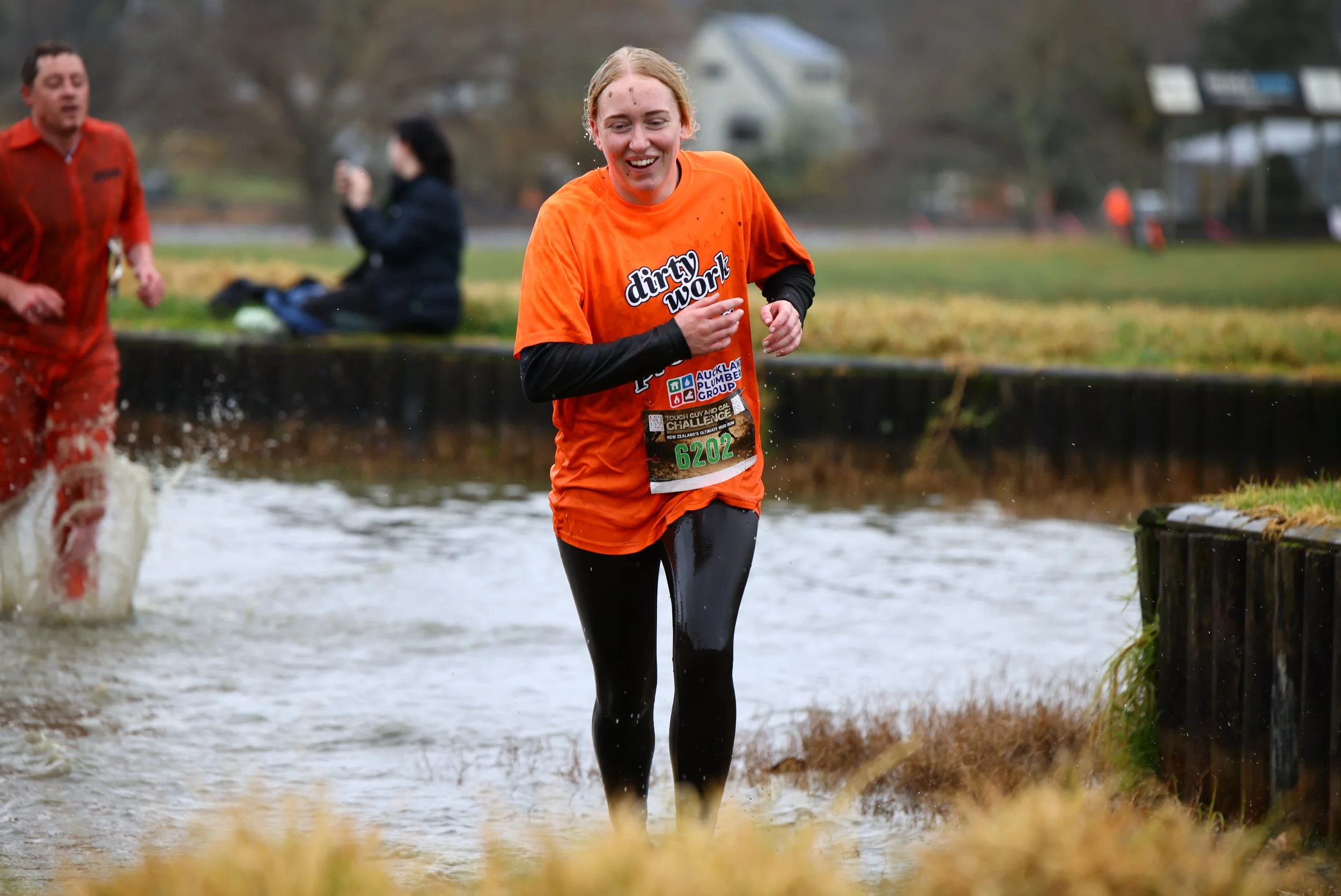 A woman running through a water crossing during a race, wearing an orange shirt with the words 'dirty work' and race bib number 6202, with other runners and trees in the background.