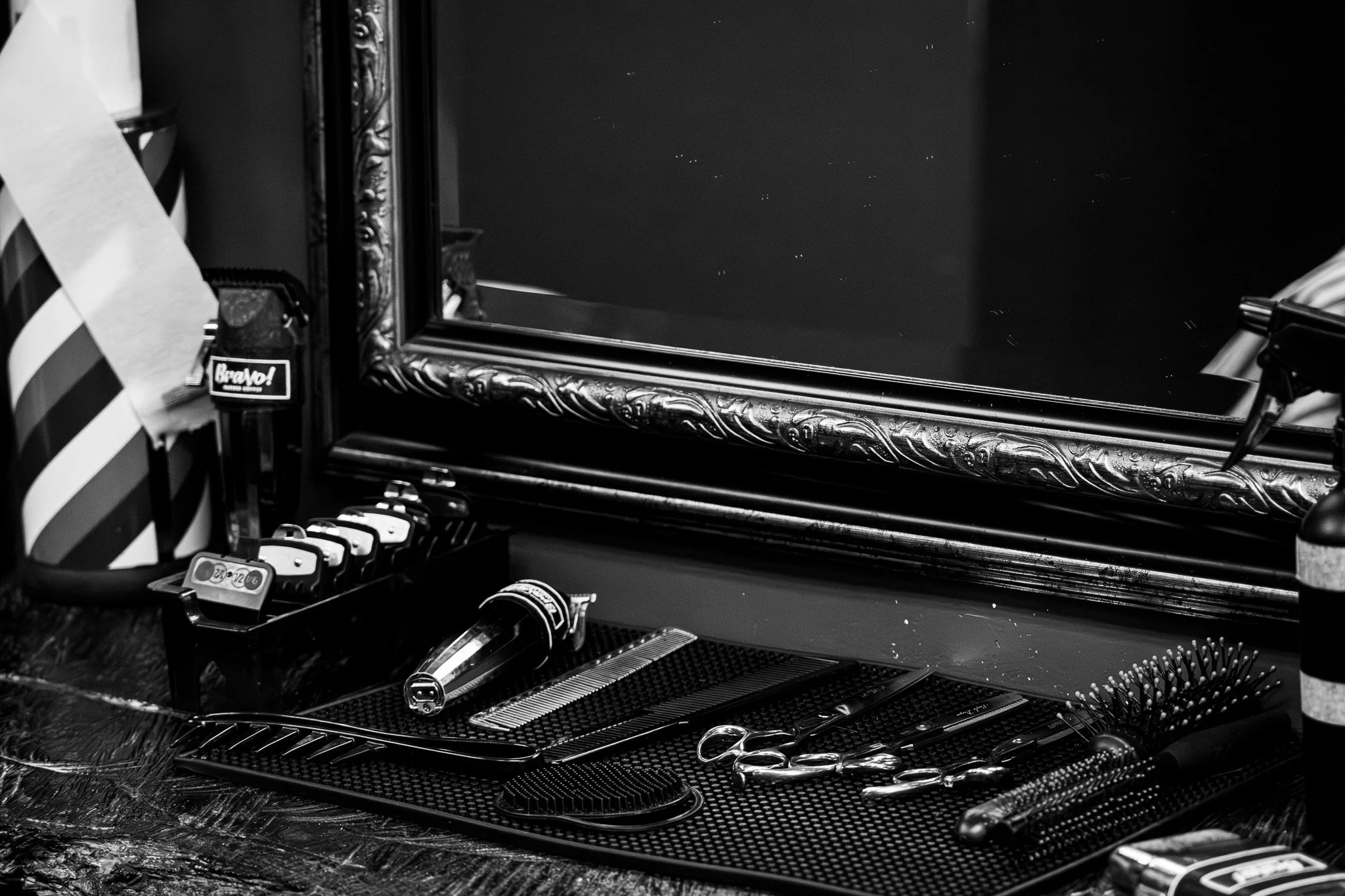 Barber shop tools including scissors, combs, and hair trimmers on a black textured surface with a large ornate mirror in the background.