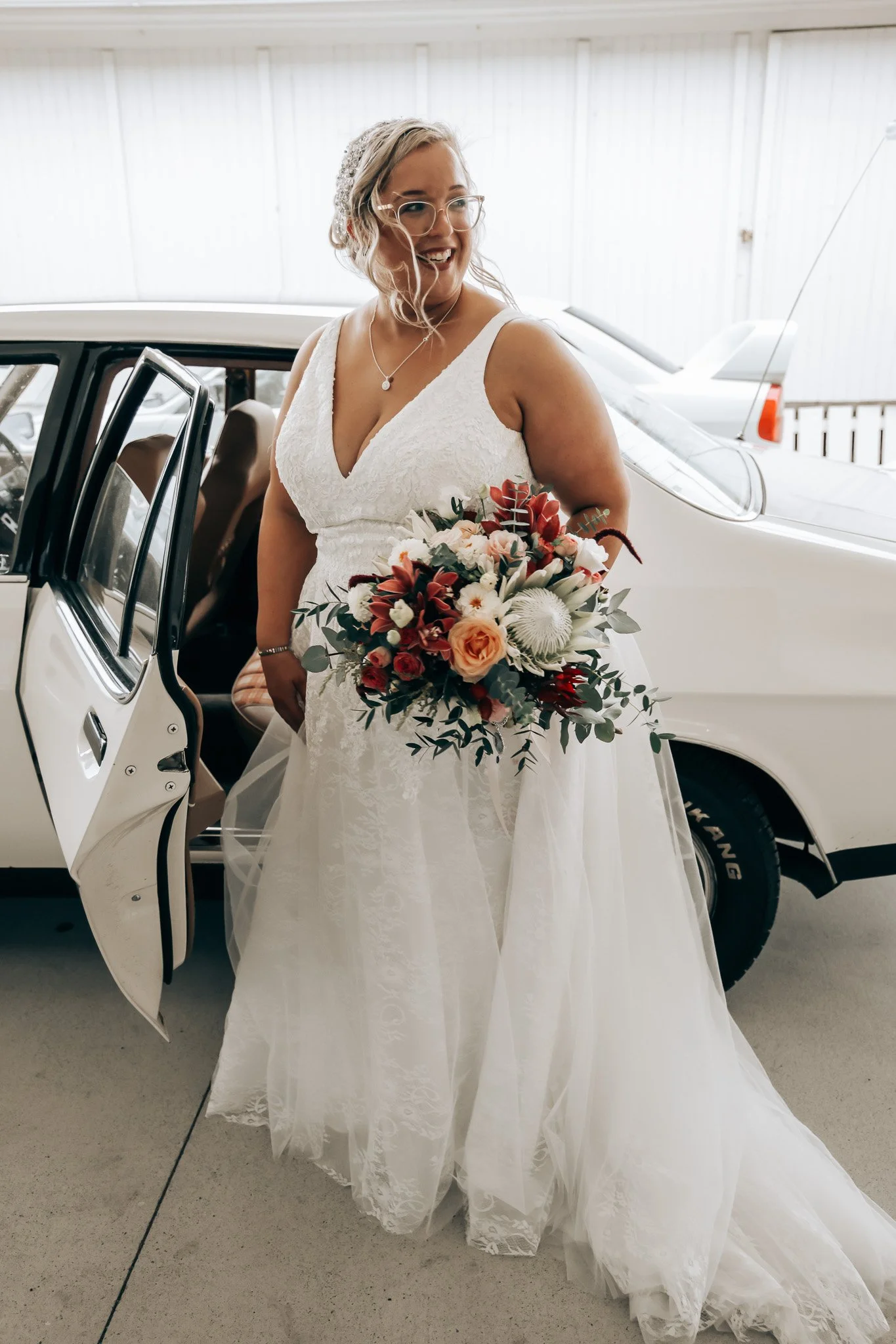 A bride in a white lace wedding gown holding a bouquet of flowers, standing next to a vintage white car with its door open, smiling indoors.