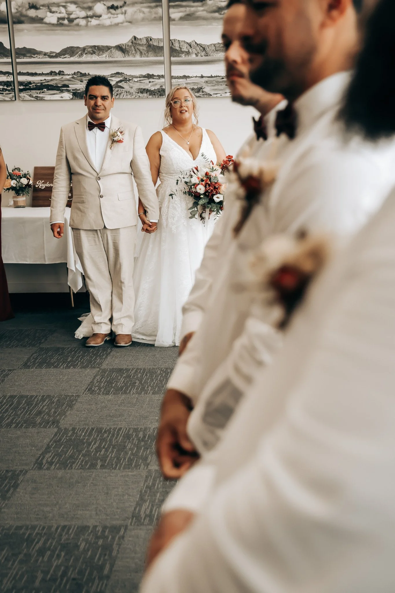 A bride and groom holding hands during a wedding ceremony, with bridesmaids in the foreground. The couple is dressed in wedding attire, standing in front of a mountain landscape painting in an indoor setting.