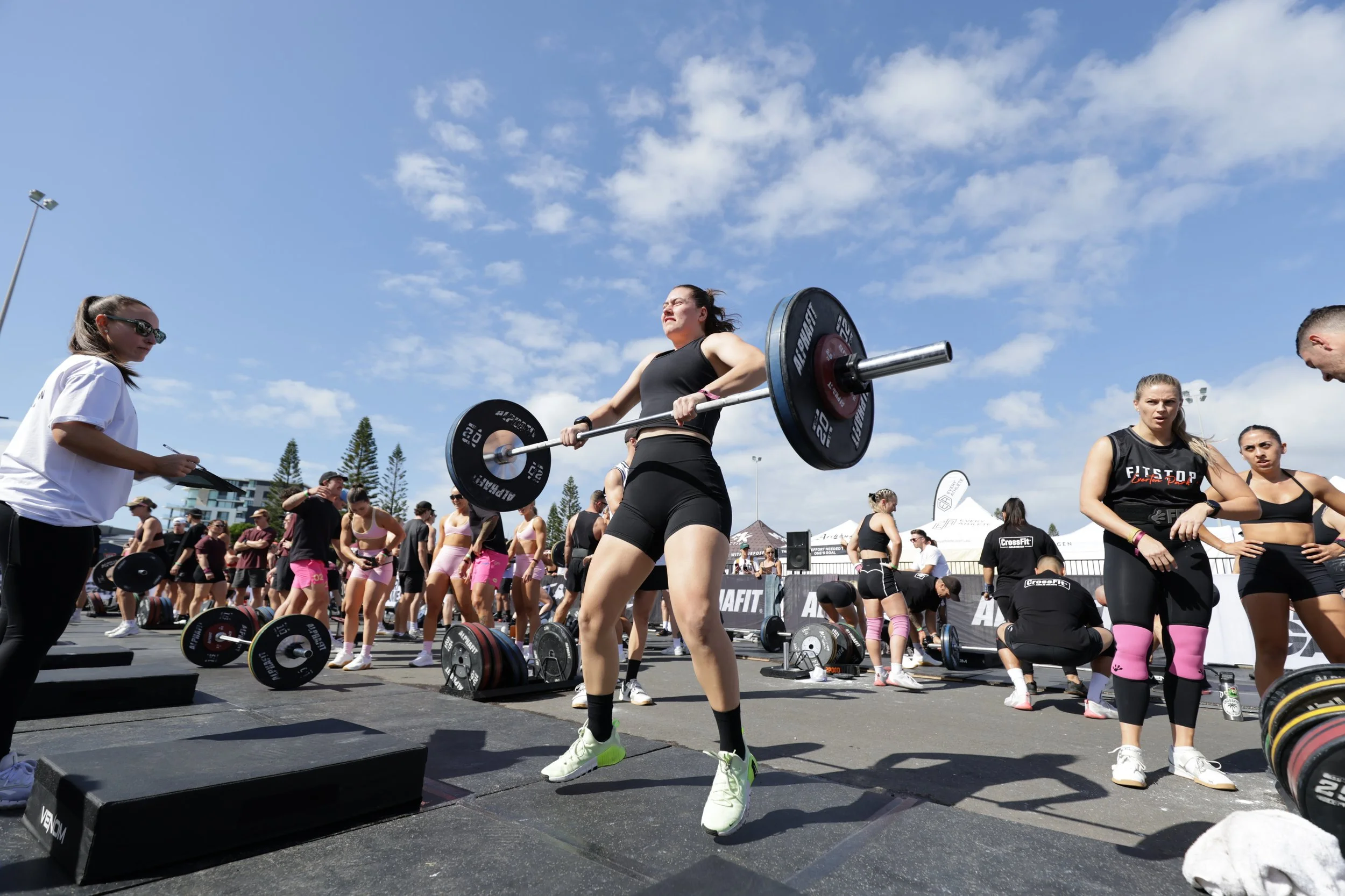 Women lifting barbells during a CrossFit outdoor event with many spectators and other athletes in the background under a partly cloudy sky.