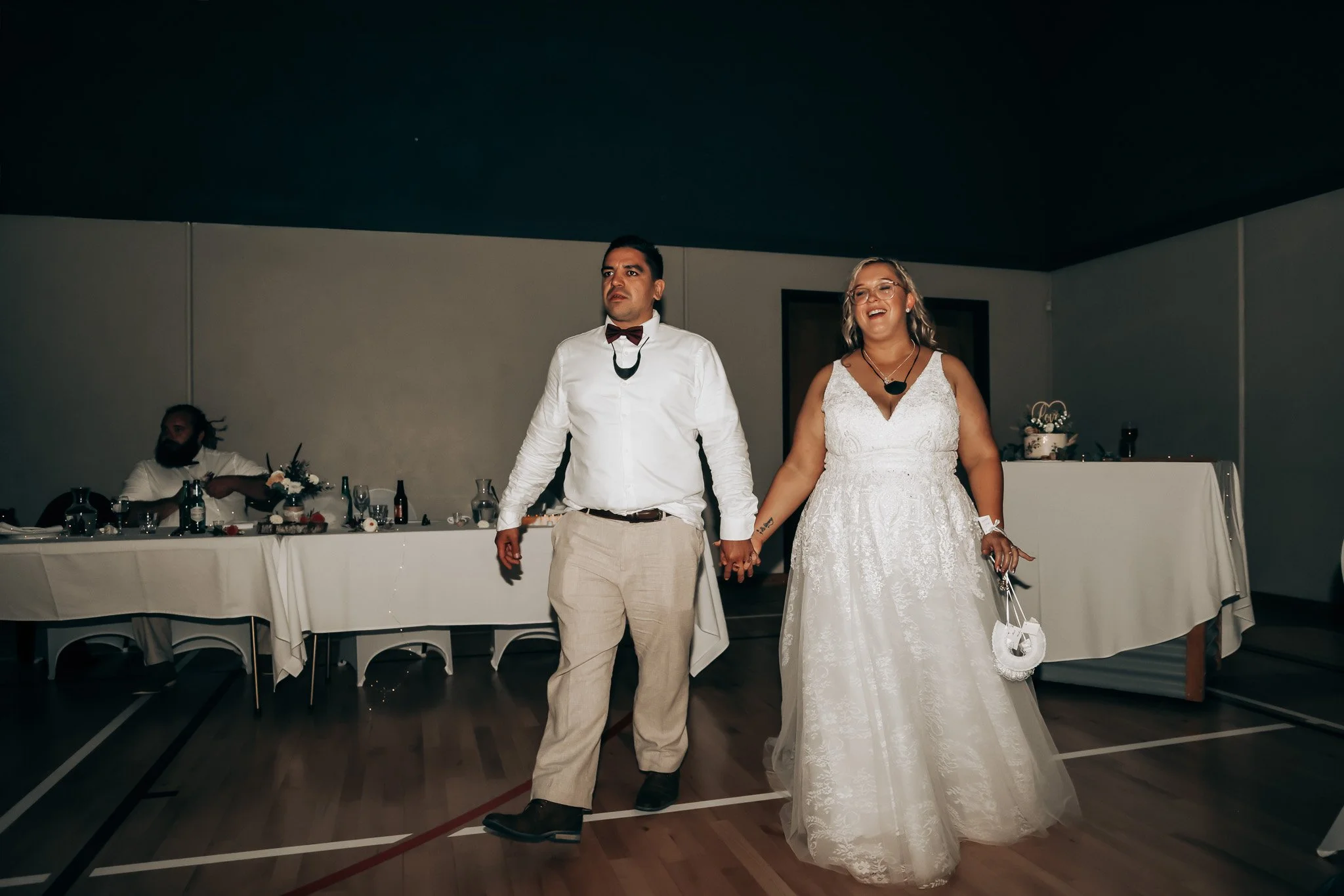 A newlywed couple holding hands and walking at their wedding reception, the bride wearing a white lace gown and the groom in a white shirt with beige pants, in a decorated hall with a table set in the background.