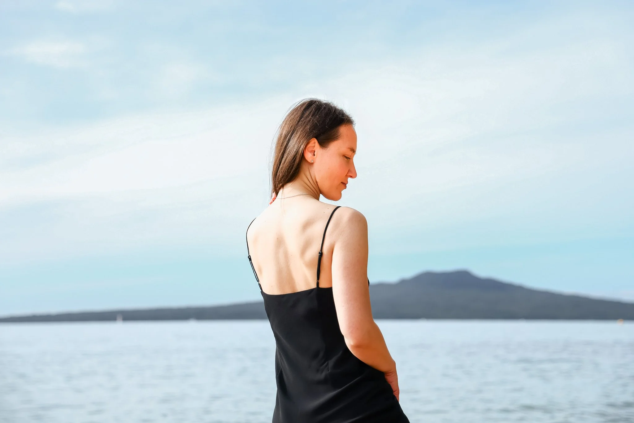 A woman with dark hair wearing a black sleeveless top, standing near water with mountains in the background, with eyes closed and looking downward.