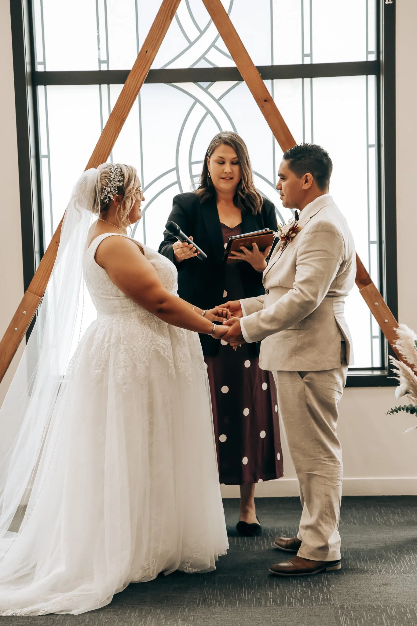 A couple holds hands during their wedding ceremony in front of an officiant, with a decorative window behind them. The bride is wearing a white wedding dress and veil, and the groom is in a cream-colored suit. They are standing on a dark carpeted flo