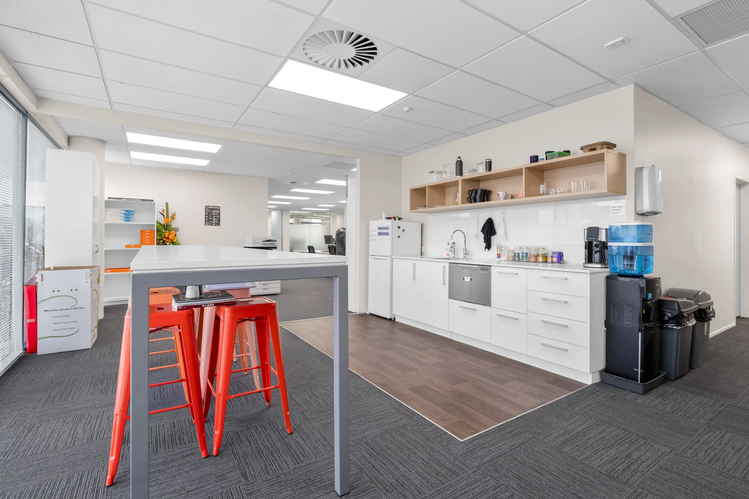 An office kitchen with white cabinets, wooden shelves holding glasses and cups, a coffee maker, water filter, and various supplies, with a gray and black carpeted floor and large windows letting in natural light.