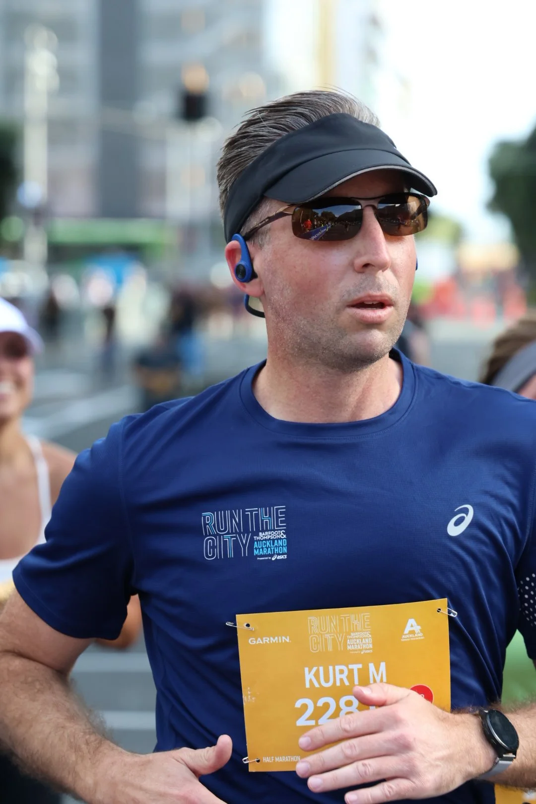 A male runner wearing sunglasses, a black visor, a blue t-shirt, and a yellow race bib with the name Kurt M participating in a marathon or race event on a city street.