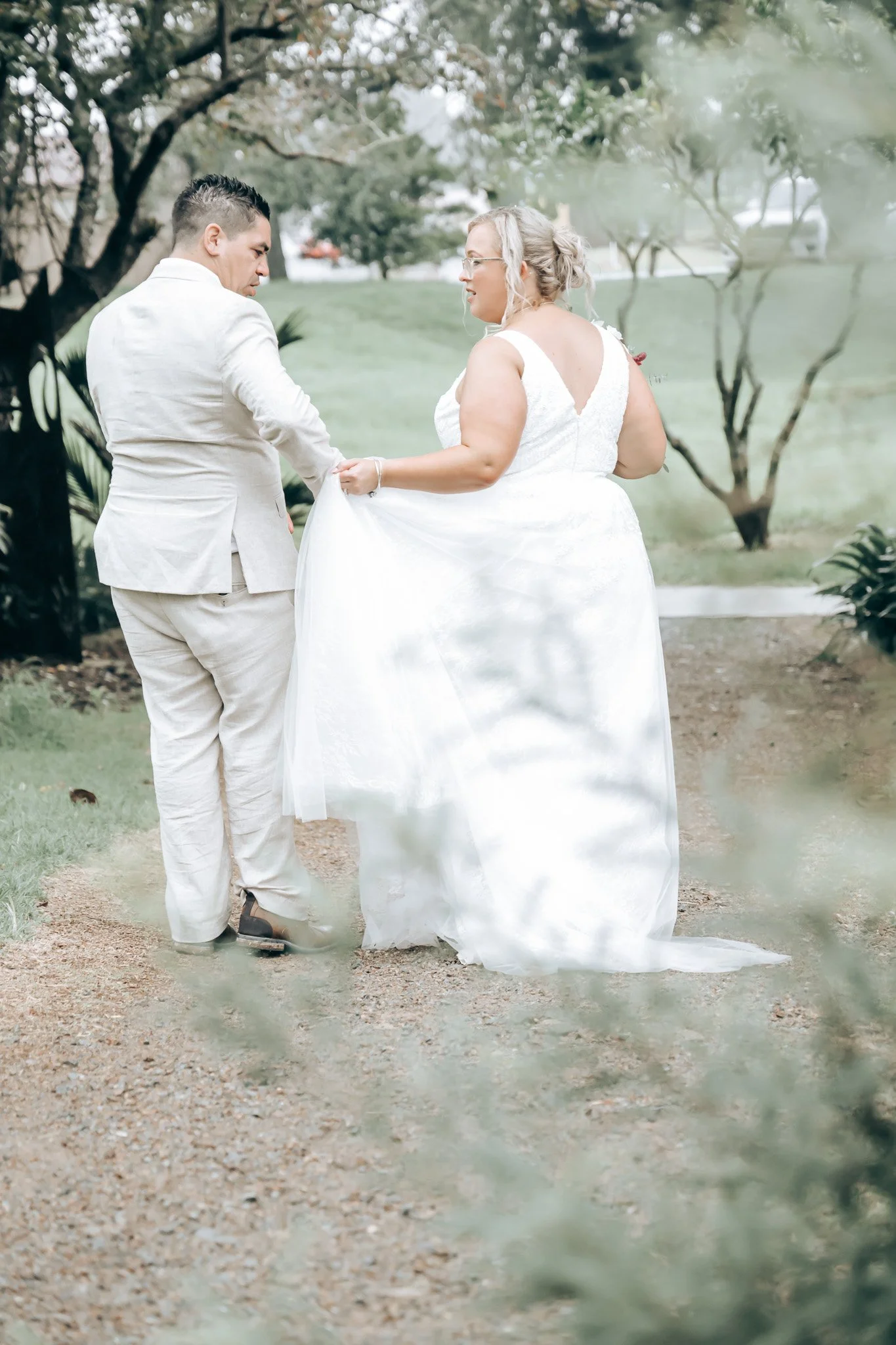 A bride in a white wedding dress holding hands with a groom in a light-colored suit in a park.