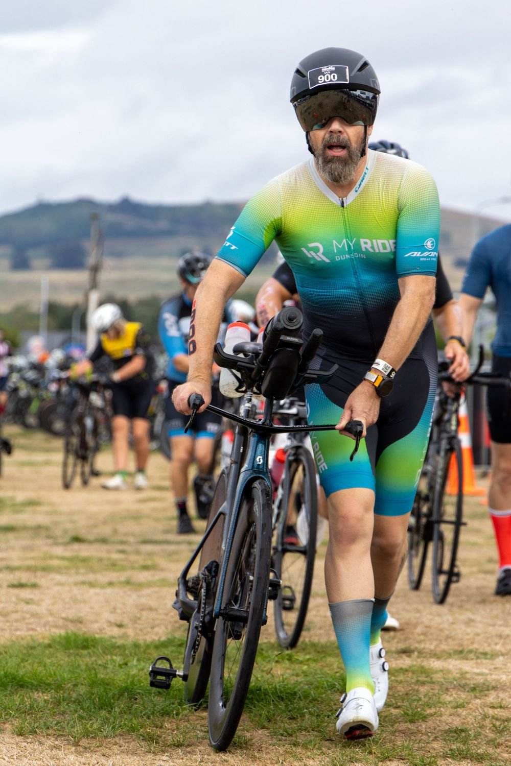 A man in colorful cycling gear, wearing a helmet and sunglasses, pushing a bike during a cycling event, with other cyclists and a partly cloudy sky in the background.
