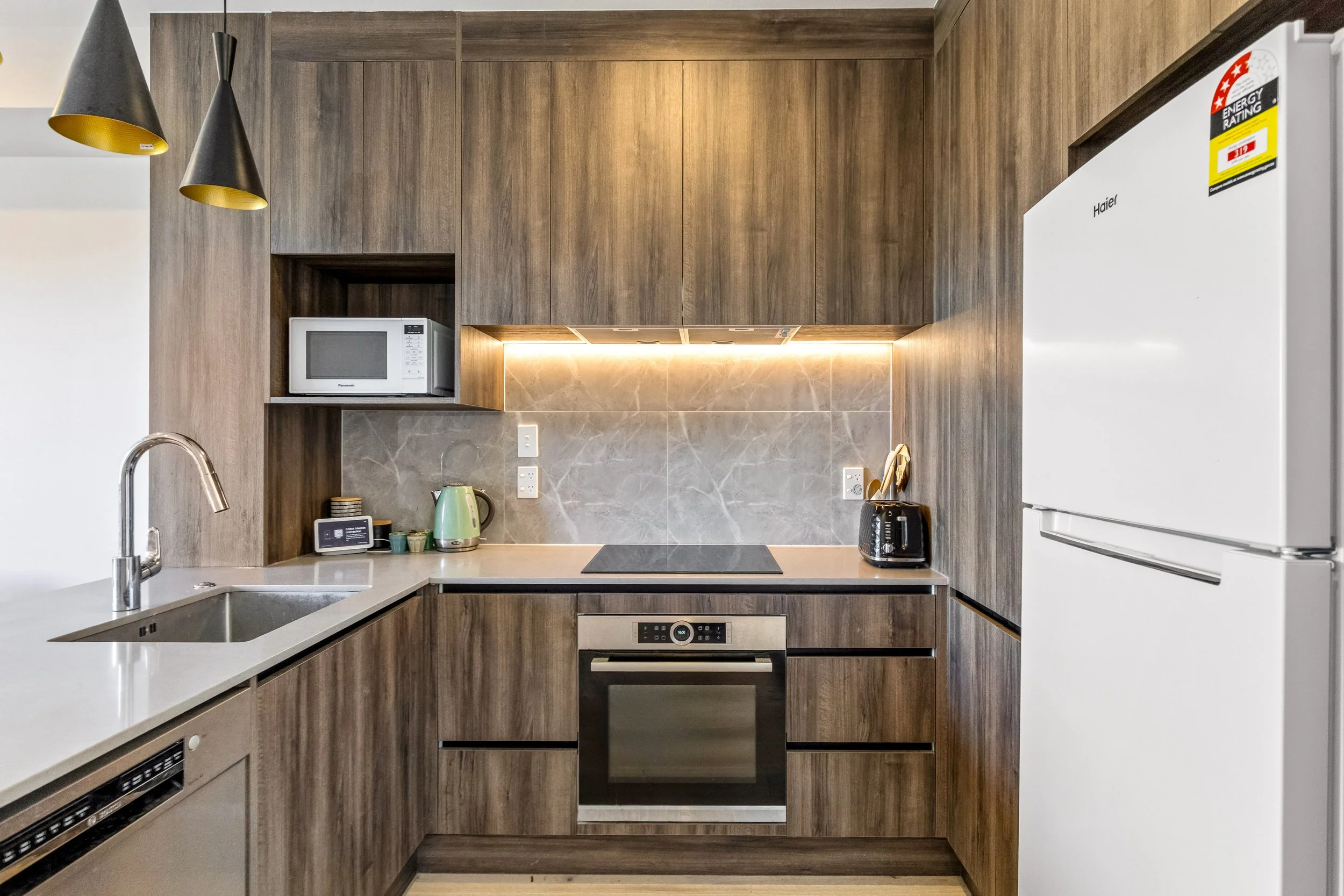 A modern kitchen with brown wood cabinetry, beige marble backsplash, and white appliances. Contains a microwave, toaster, electric kettle, and oven, with pendant lights hanging from the ceiling.