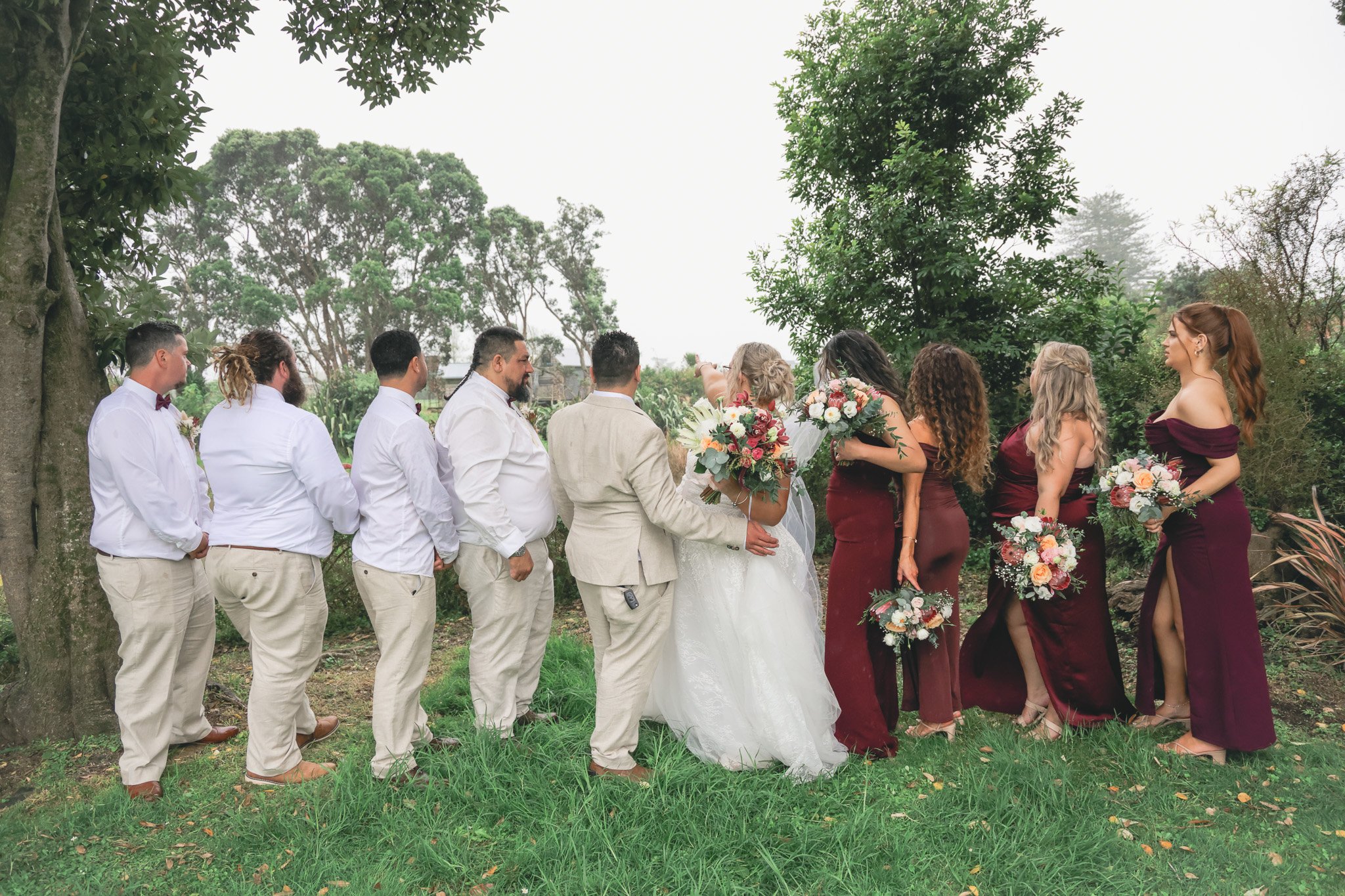 A wedding ceremony outdoors with a bride and groom exchanging vows, surrounded by bridesmaids and groomsmen. The scene is set in a green, wooded area with trees and grass.