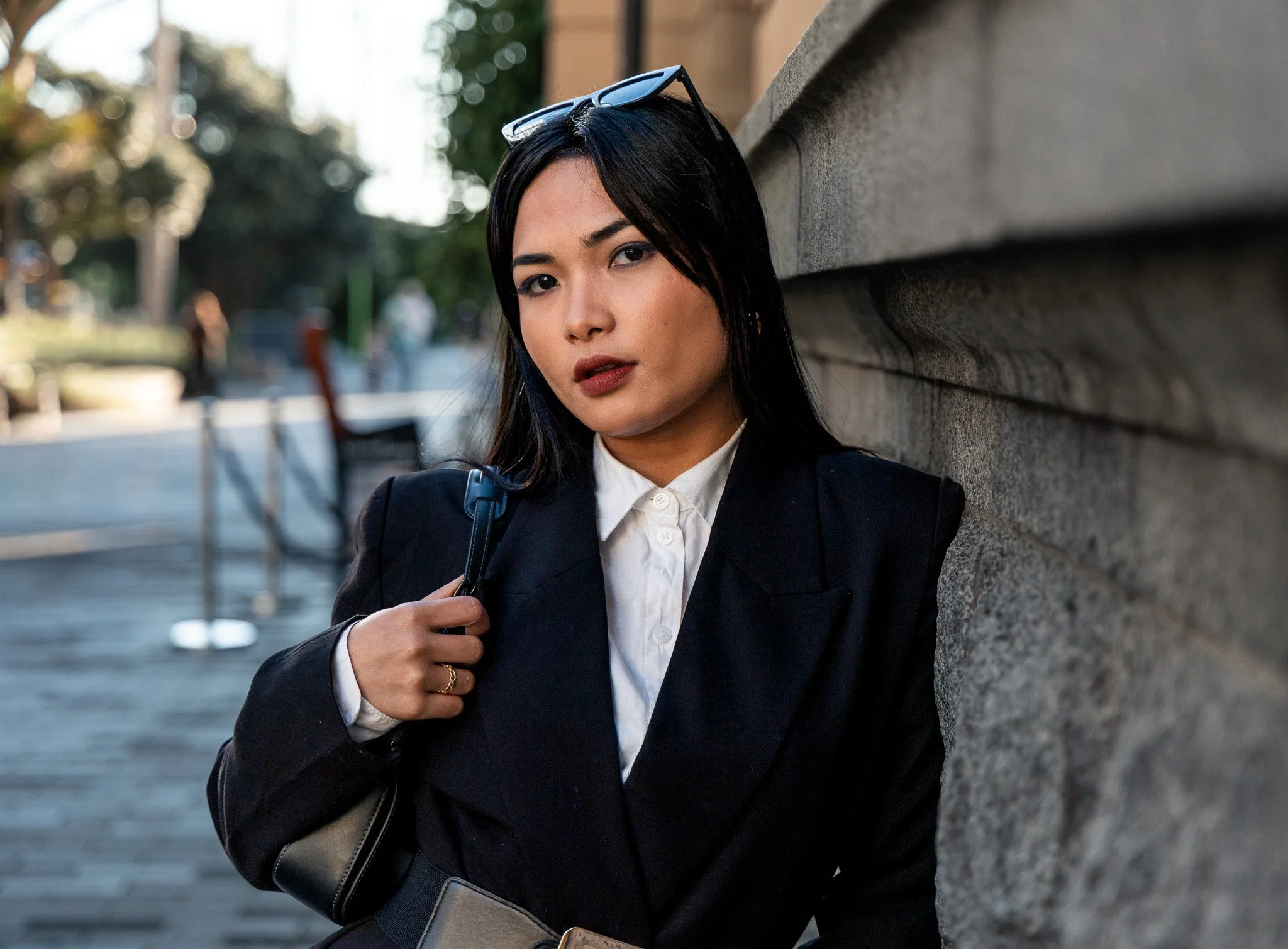 A woman with dark hair wearing glasses on her head, dressed in a navy blazer and white shirt, standing against a stone wall outdoors.