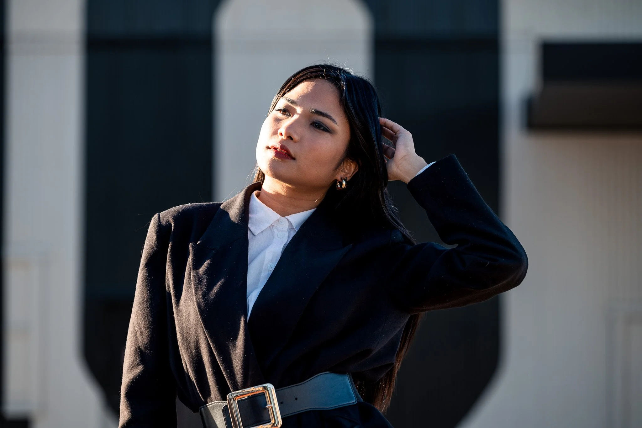 A young woman with dark hair wearing a black blazer and white shirt, posing outdoors with her hand behind her head, standing in front of a modern building with dark and light panels.