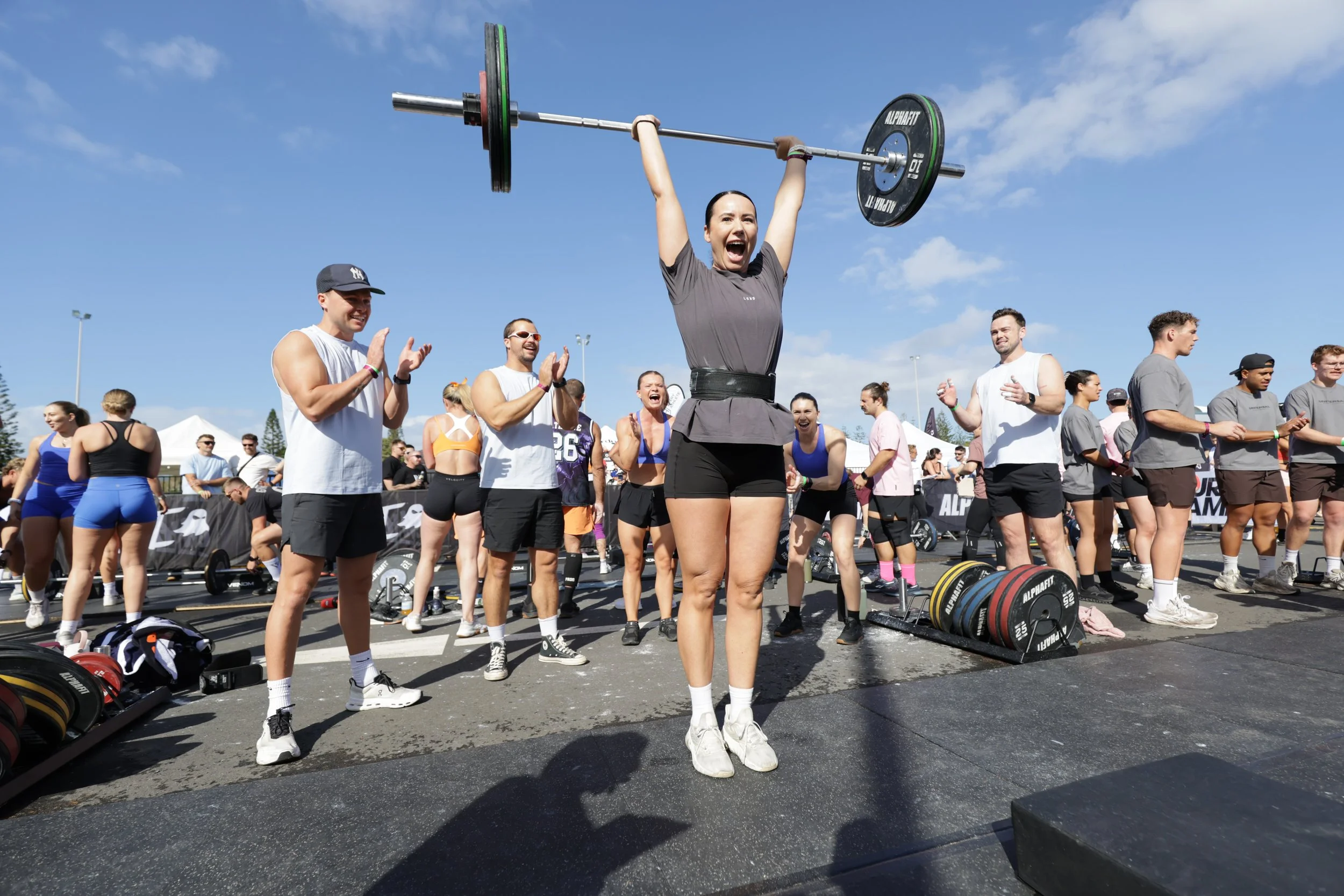 A woman lifts a barbell overhead at a fitness event, surrounded by a crowd of people clapping and celebrating outdoors on a bright, sunny day.