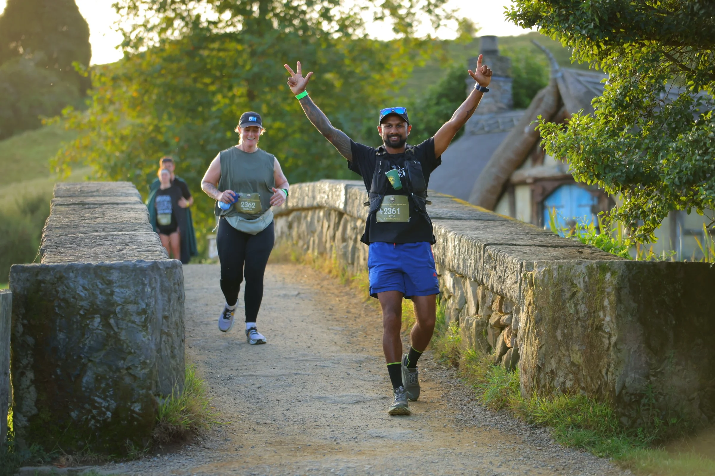 A group of people participating in a running event outdoors on a stone path with green trees and cottages in the background, with one man in front raising his arms in victory.