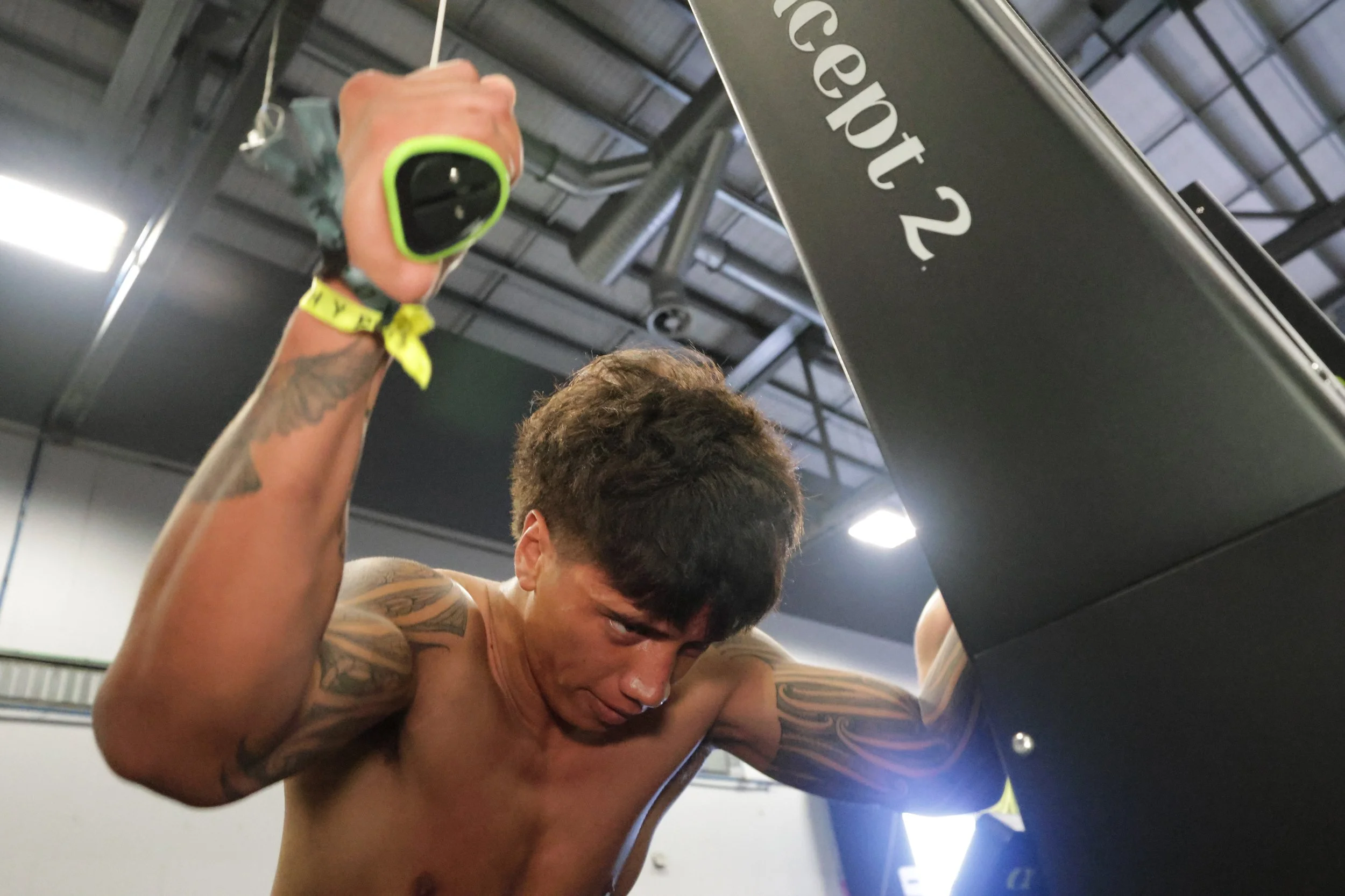 A shirtless man with tattoos on his arms and chest, leaning forward with his head down, appears to be exercising or competing in a gym, with gym equipment and industrial ceiling visible in the background.