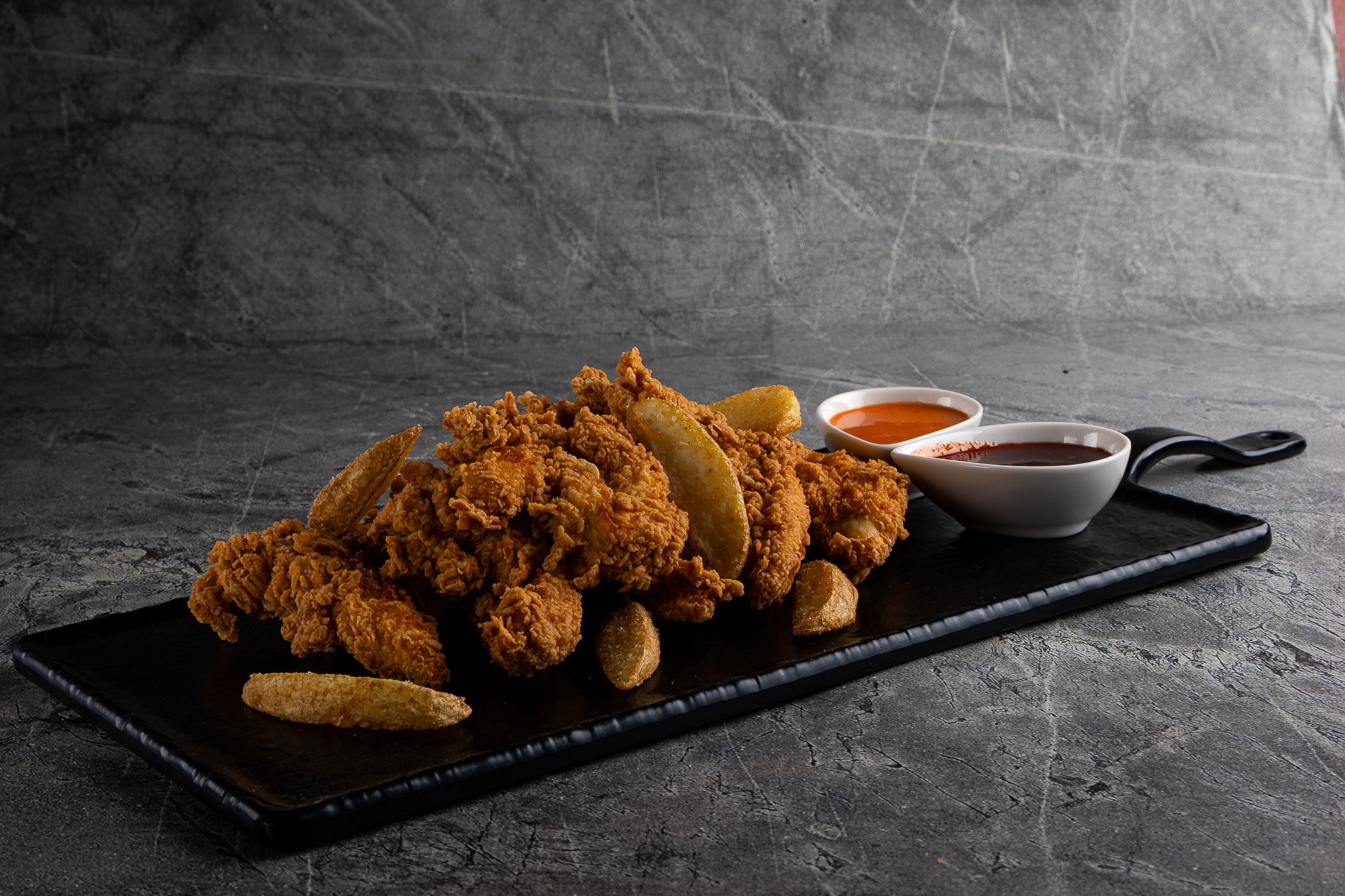 A black rectangular serving plate with fried chicken pieces, potato wedges, and potato fries, accompanied by two small bowls of dipping sauces.