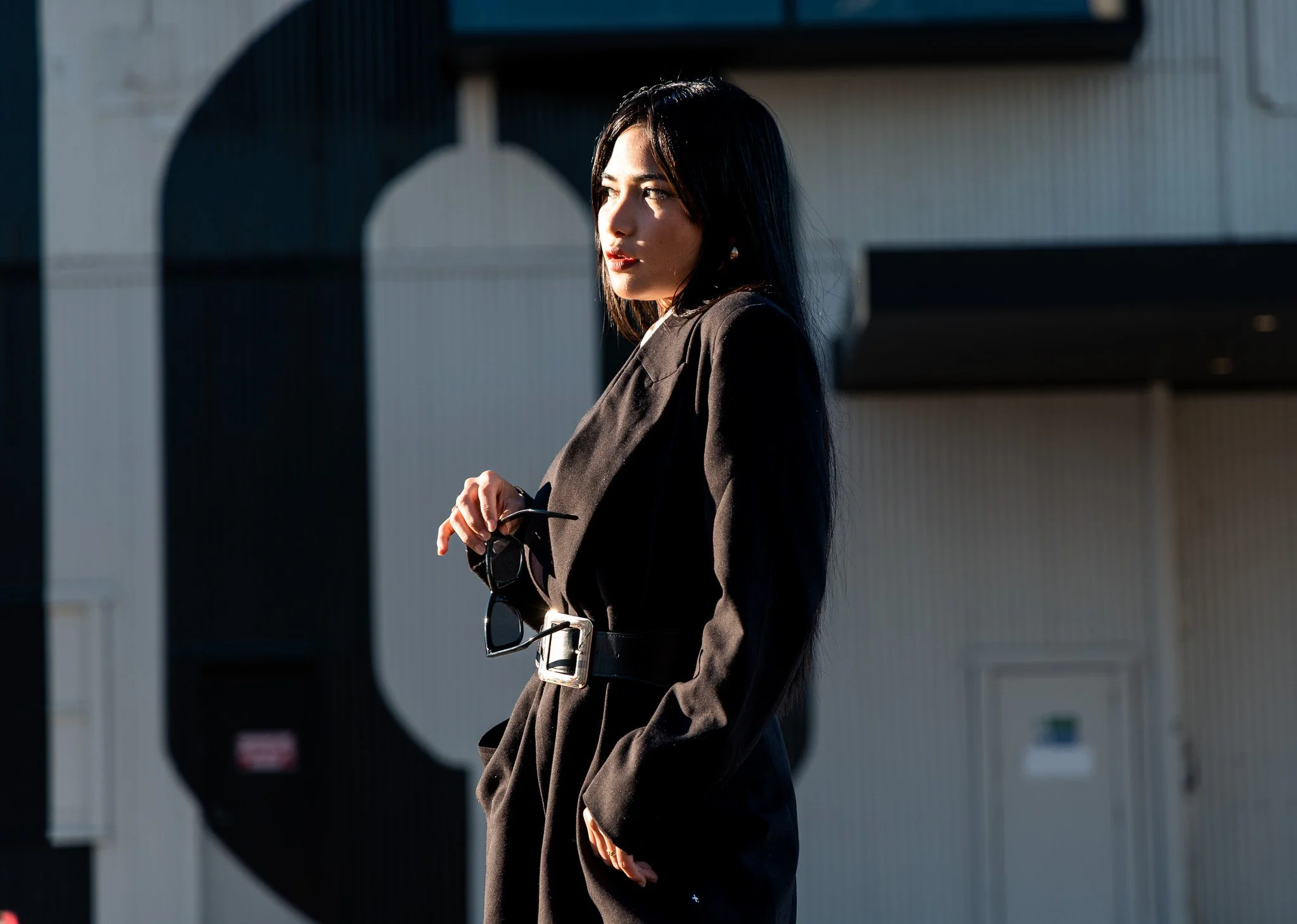 A woman with dark hair, dressed in a black blazer, stands outdoors with sunlight illuminating her face. She is holding sunglasses and gazing to the side with a thoughtful expression, with a modern building in the background.
