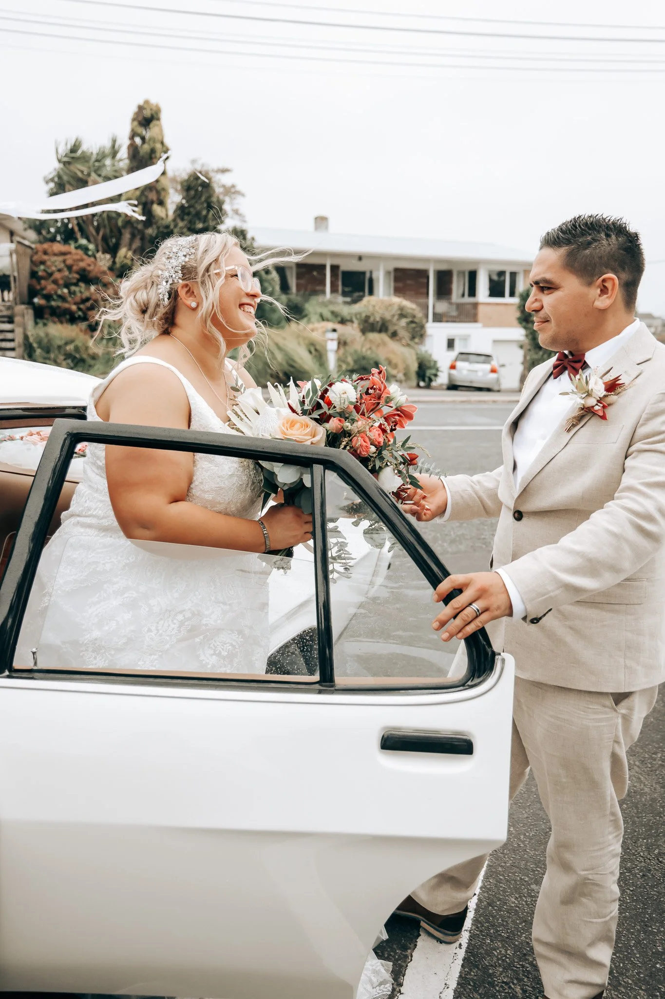A bride and groom exchanging vows outside near a white vintage car, with the bride holding a bouquet of flowers and the groom wearing a light-colored suit with a bow tie. They are smiling at each other on a cloudy day, with houses and a parked car in