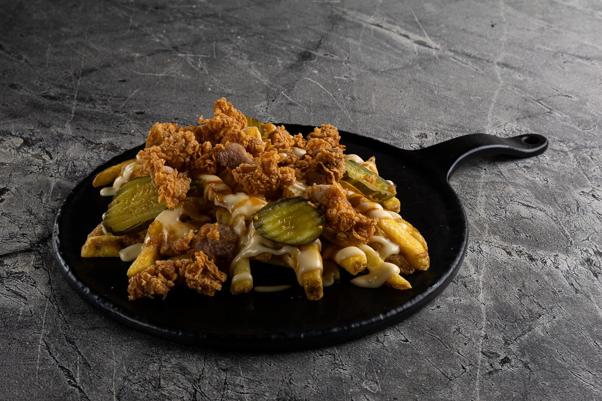 A black plate of loaded fries topped with fried chicken bites, pickles, and drizzled with mayonnaise or ranch dressing on a dark textured surface.