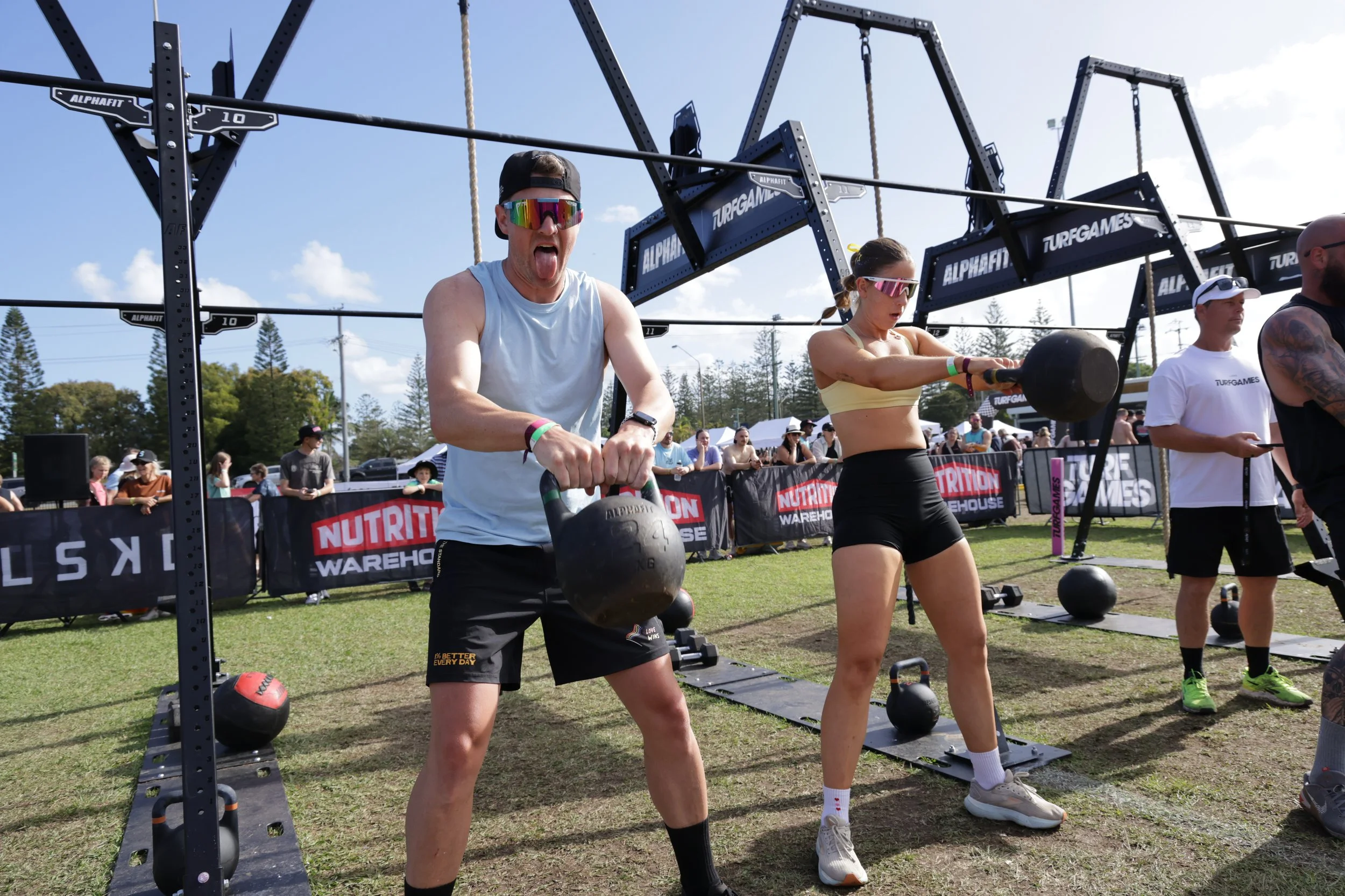 People participating in an outdoor fitness event, lifting kettlebells, with a large fitness rig and crowd in the background.
