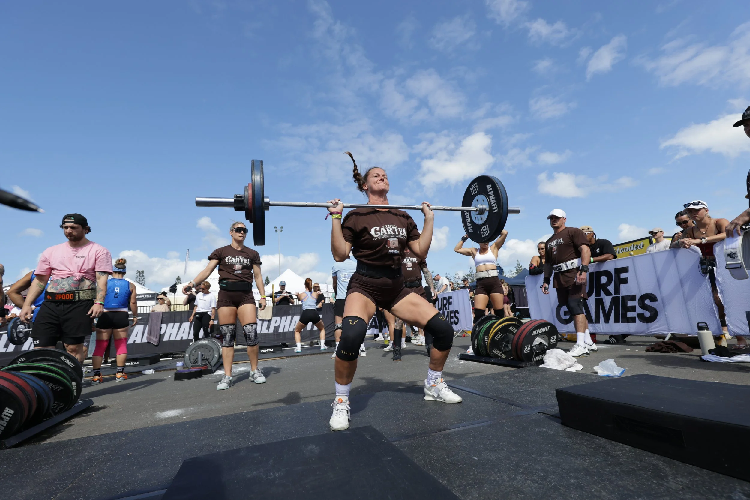 Woman lifting a barbell at an outdoor fitness competition with spectators and equipment around her under a partly cloudy sky.