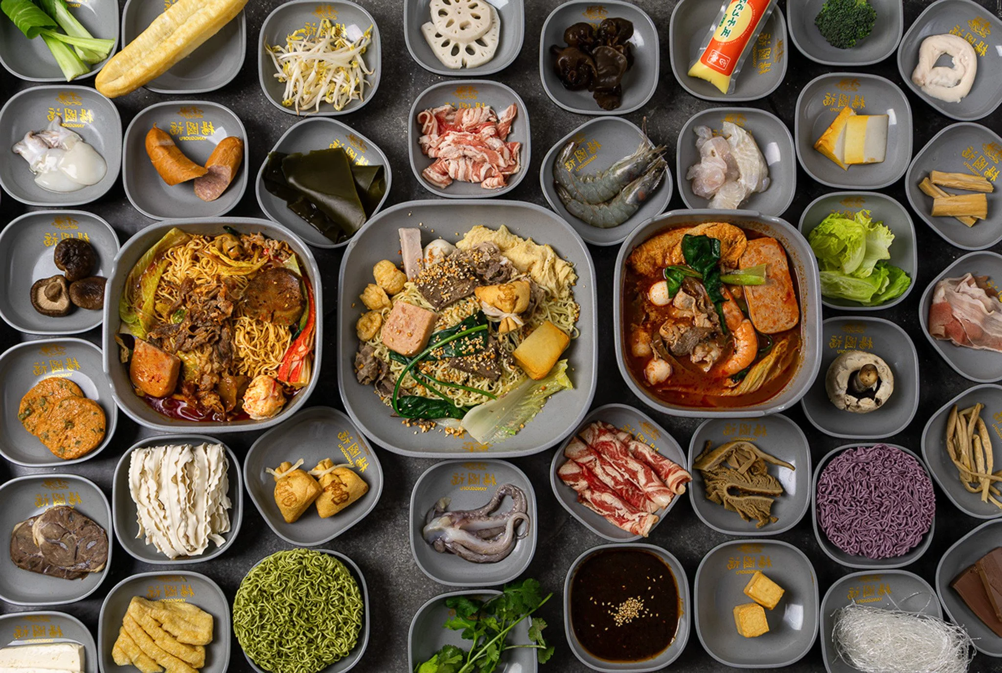 A variety of ingredients for hot pot, including vegetables, meats, seafood, and noodles arranged in small bowls on a dark table.