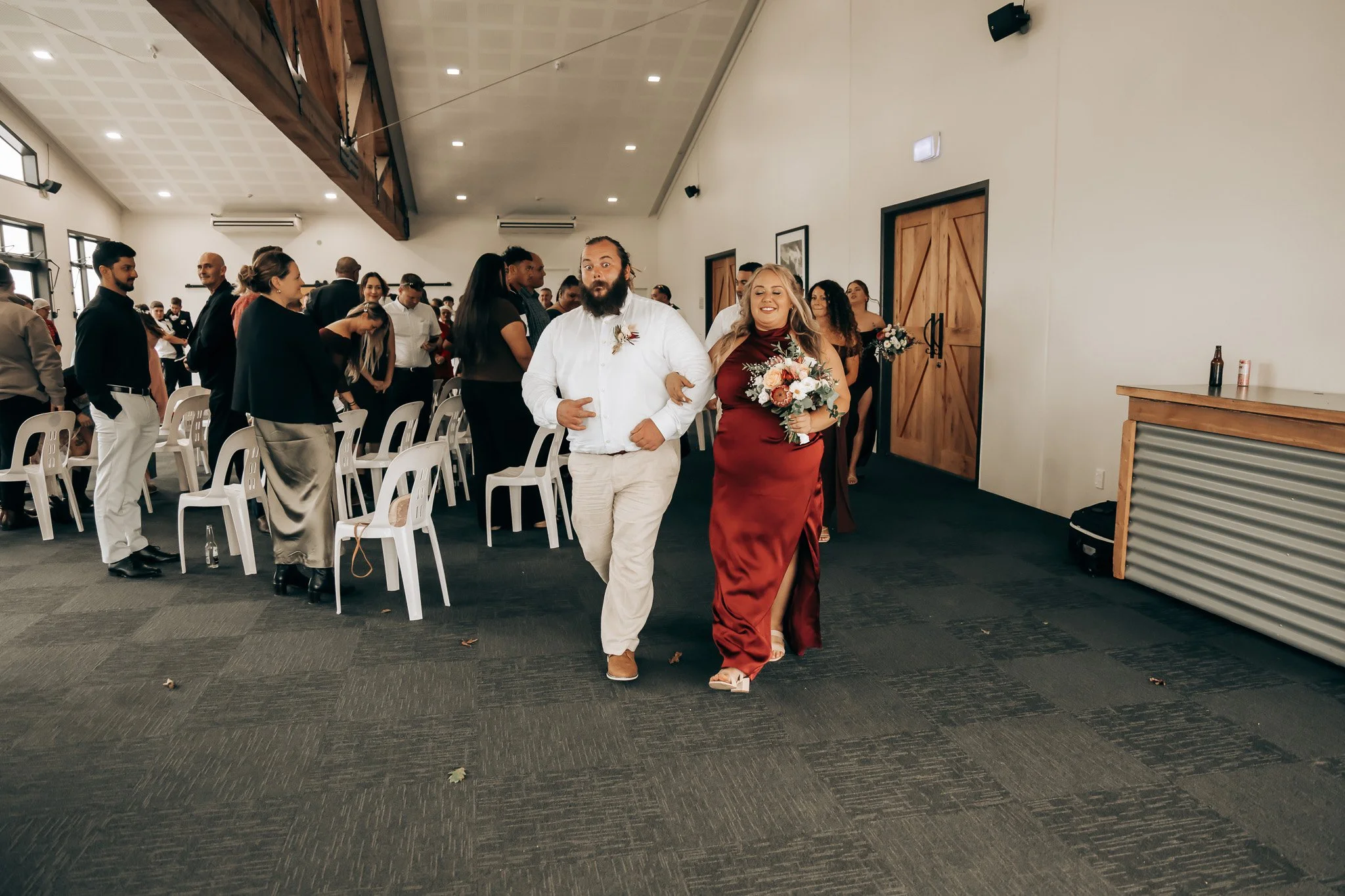 A bride in a burgundy wedding dress walking arm in arm with a groom in white attire, surrounded by wedding guests in a decorated indoor venue.