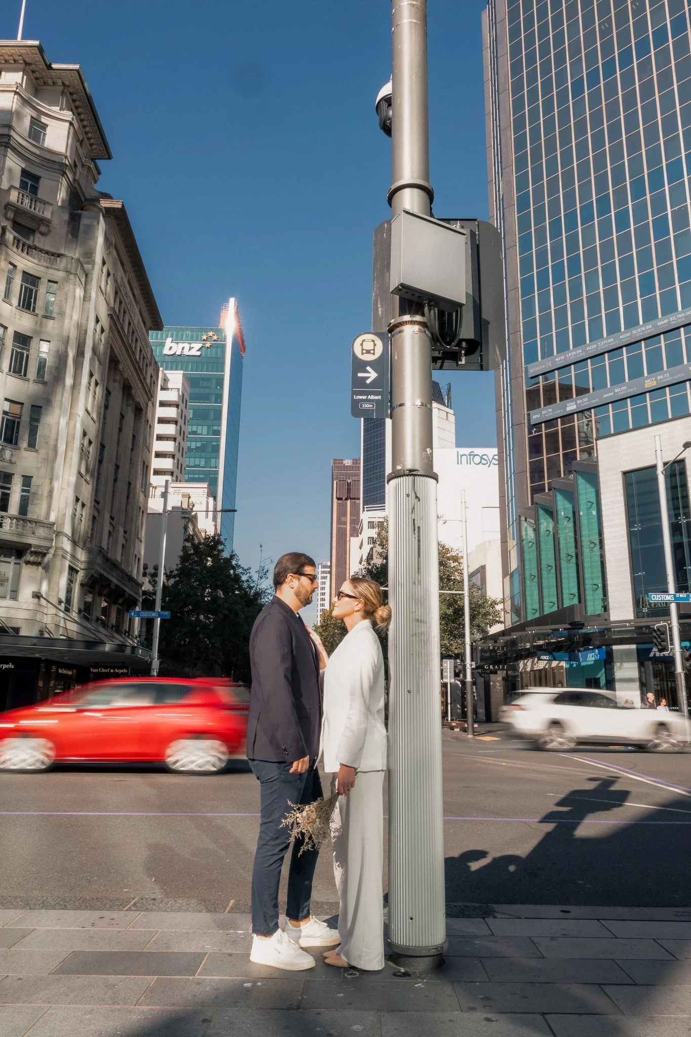 A man and a woman in formal attire standing face to face on a city street, underneath a street lamp, with some cars passing by and tall buildings in the background.