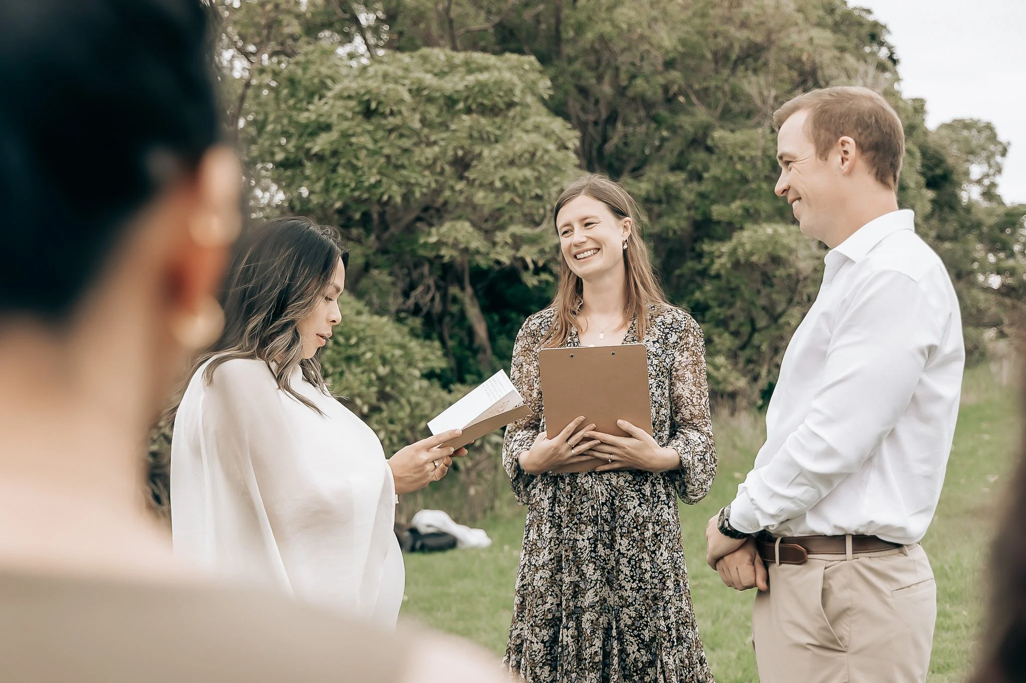 A group of people participating in an outdoor ceremony or gathering, with one woman holding a book and a clipboard, smiling, and others listening attentively, set in a park with trees in the background.