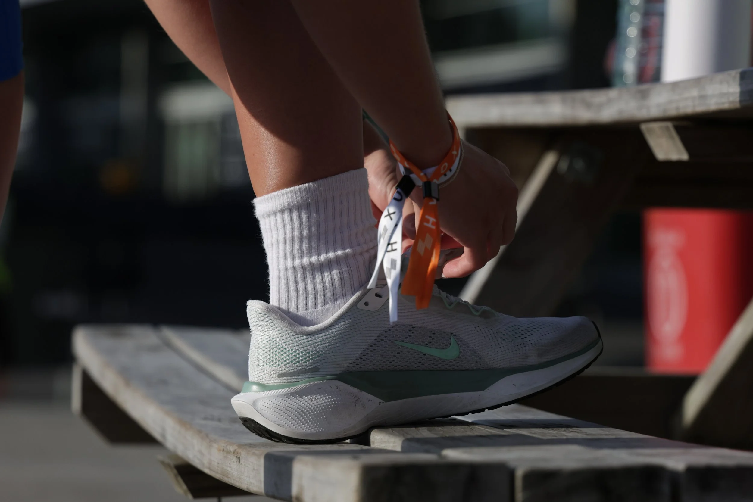 Close-up of a person's foot wearing a white running shoe with a light green swoosh and white sock, tying shoelaces while standing on a wooden bench outdoors.
