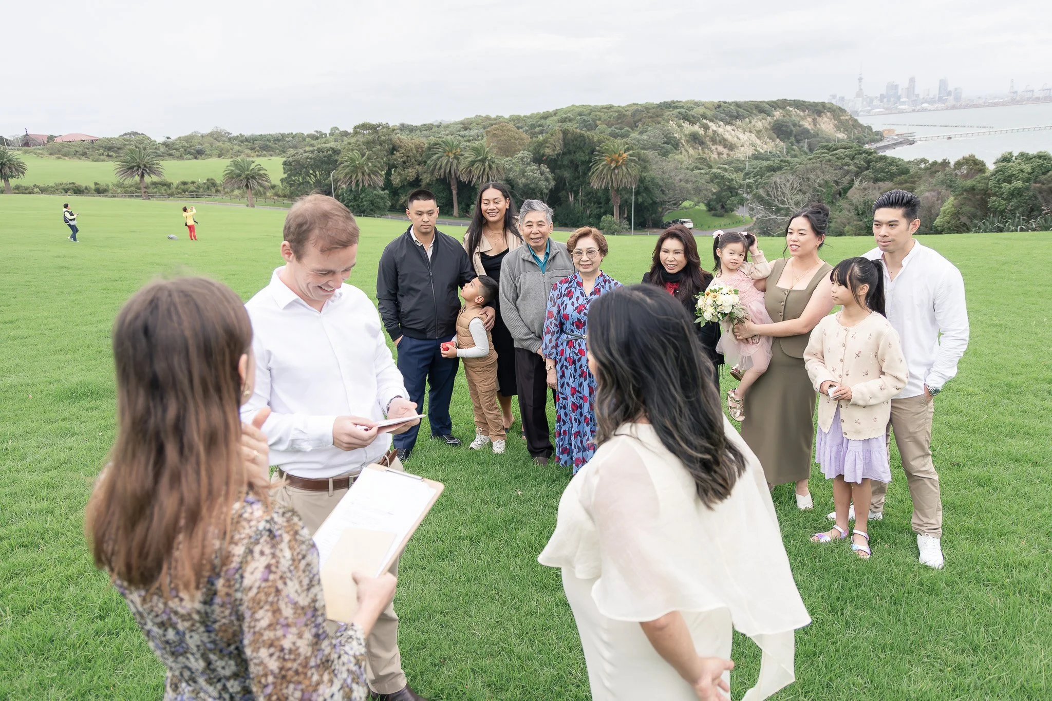 A group of people gathered outdoors on a grassy field with trees and hills in the background, engaging in a ceremony or celebration, with some individuals holding flowers, and others taking photos in the distance.