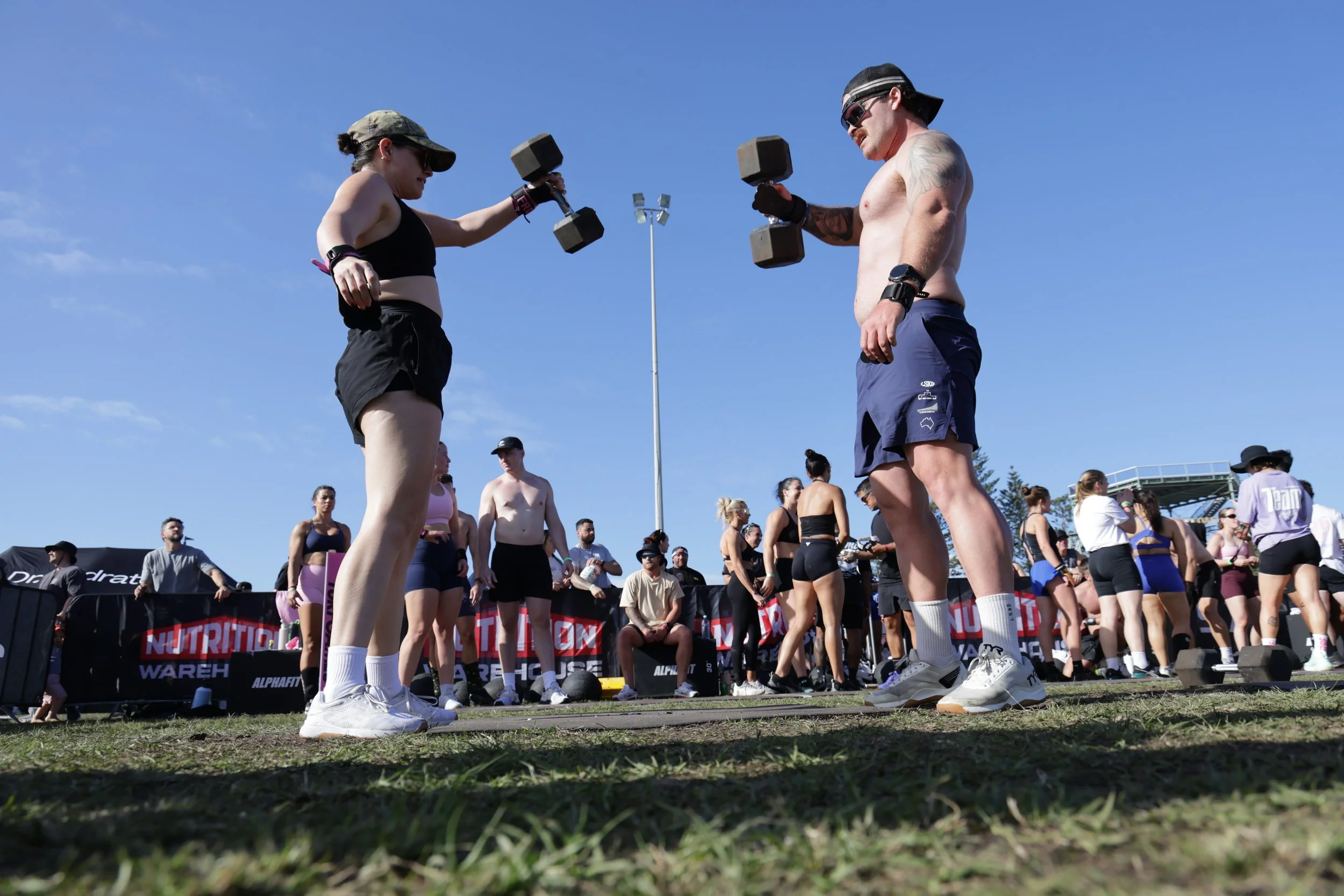 A group of people at an outdoor fitness event, with two individuals in the foreground lifting dumbbells while others stand and watch, under a clear blue sky.
