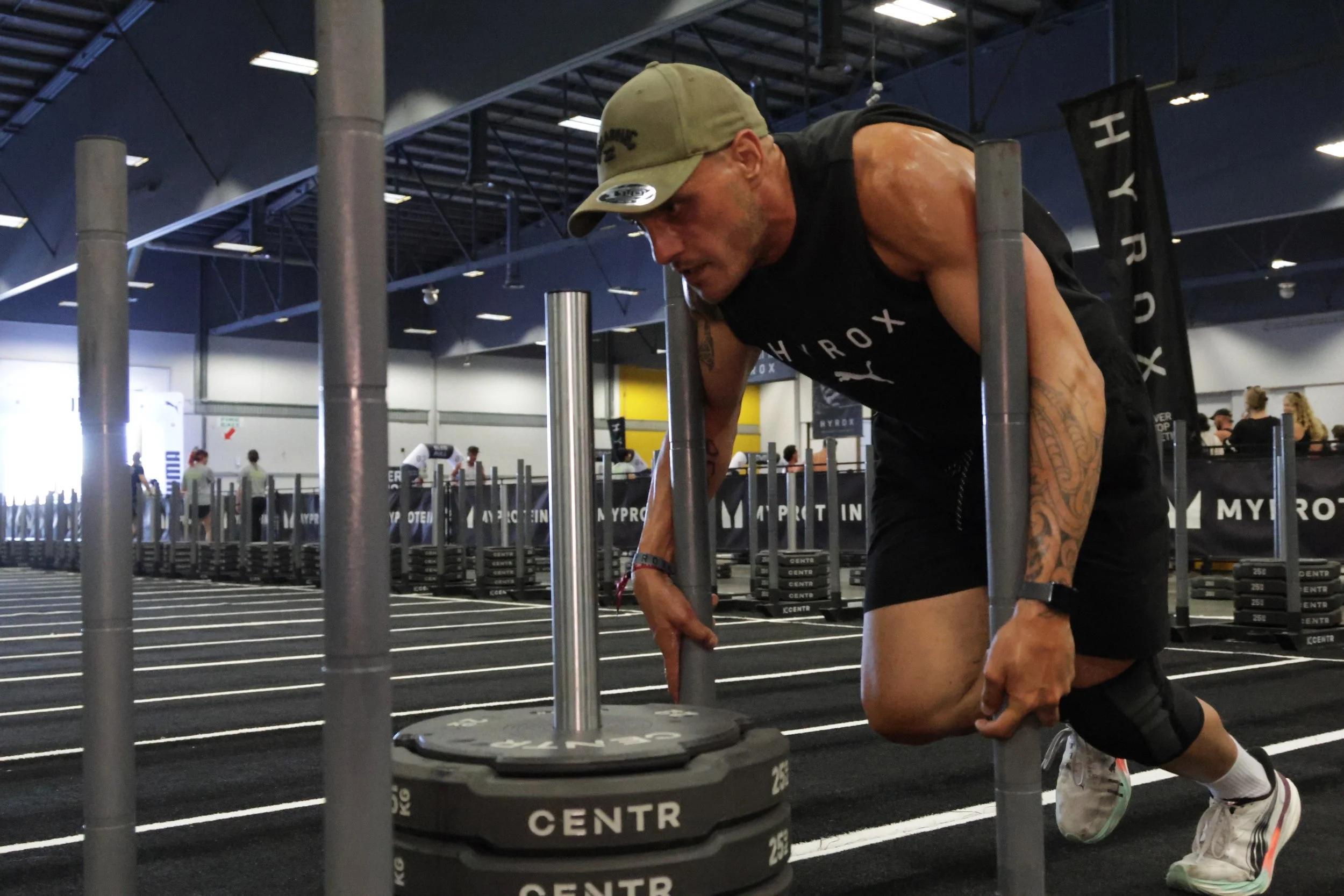 A man wearing a khaki baseball cap, black tank top, and shorts is kneeling on one knee on a gym floor, preparing for a workout. The gym has black flooring with white lines and weights stacked nearby. Several people are seen in the background, with a 
