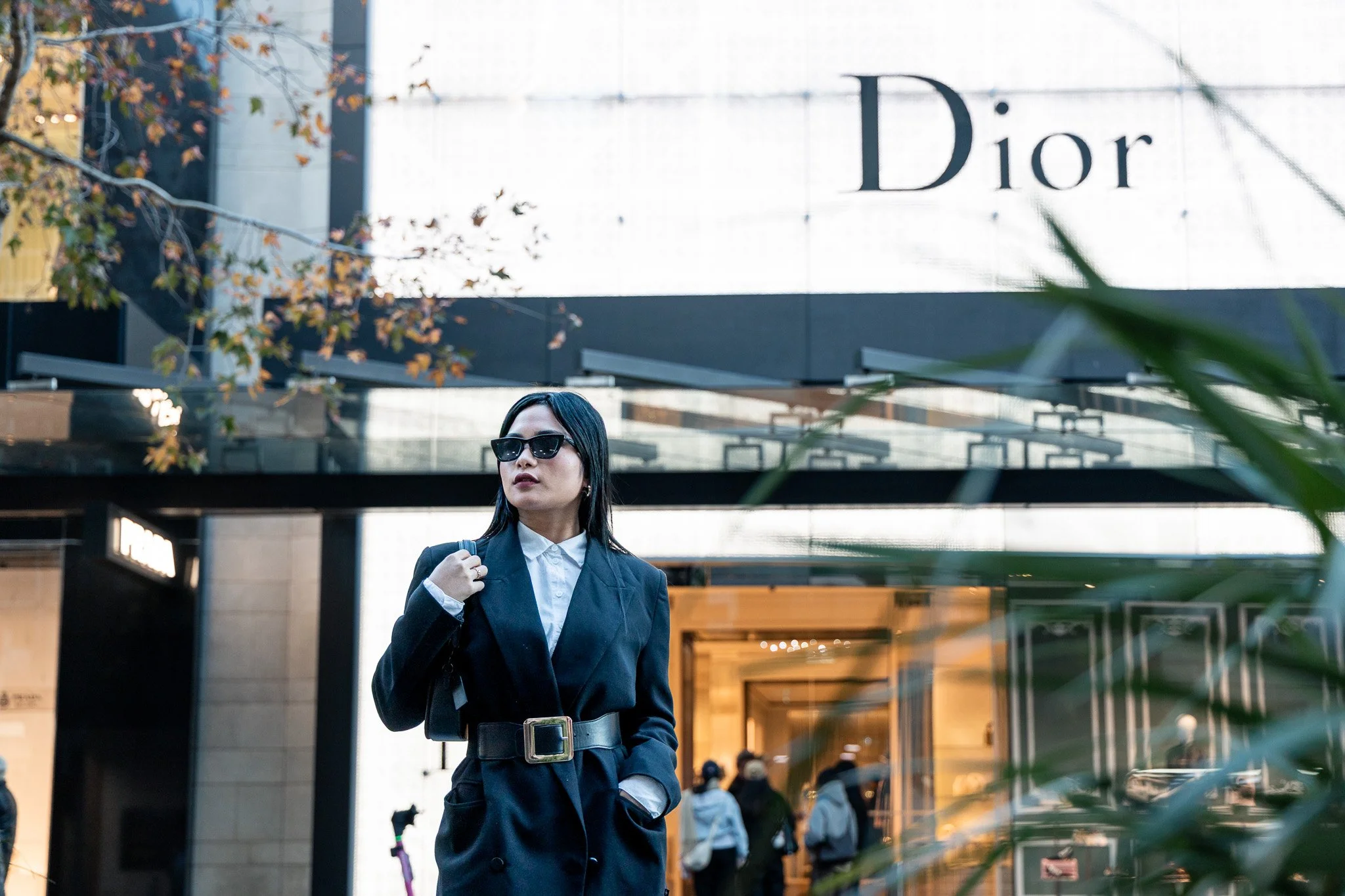 A woman in a dark suit and sunglasses walking outside a Dior store, with a building and people visible in the background.