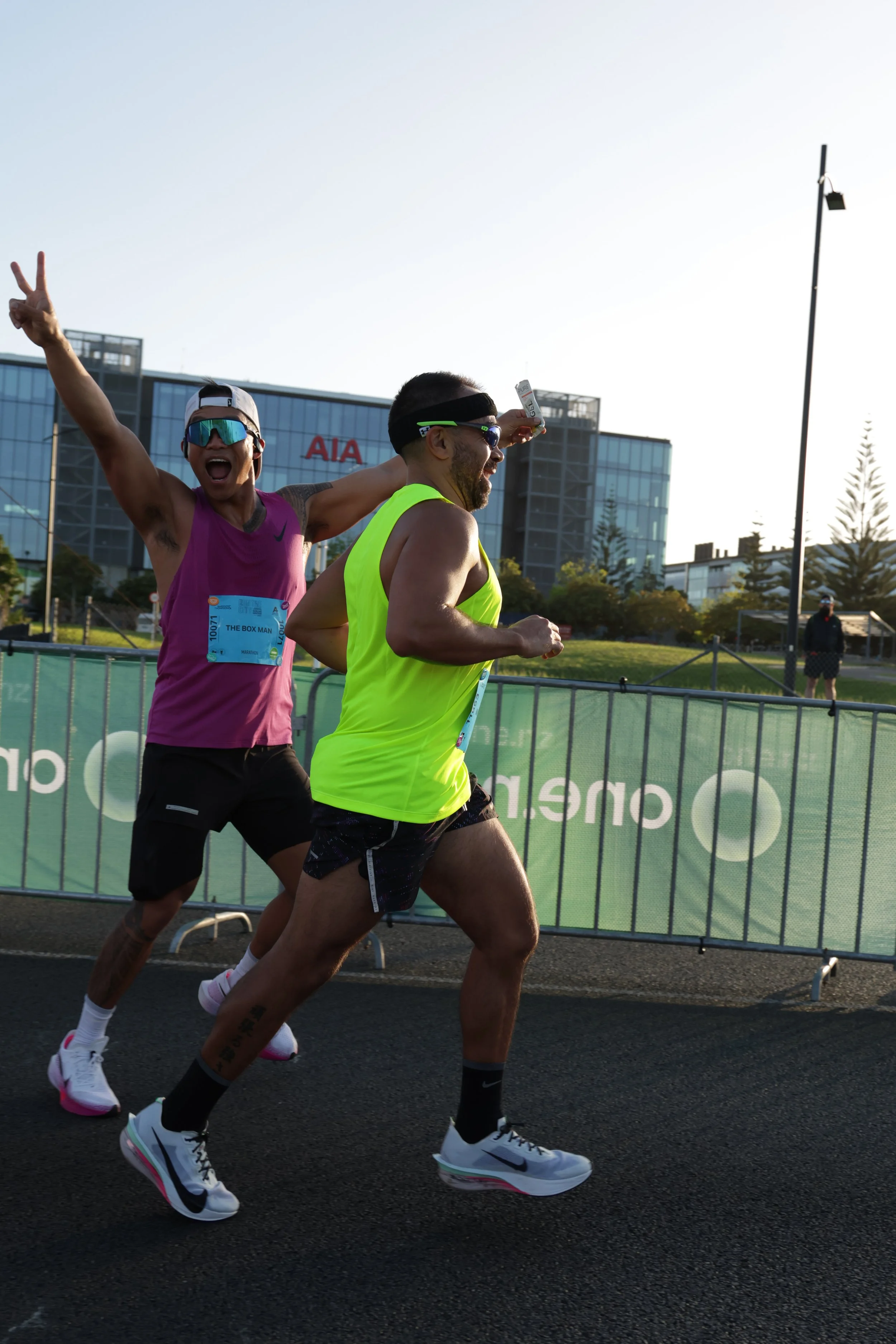 Two male runners participating in a race, one wearing a bright yellow tank top, the other in a purple shirt, with a cityscape background including modern buildings and a green race barrier.