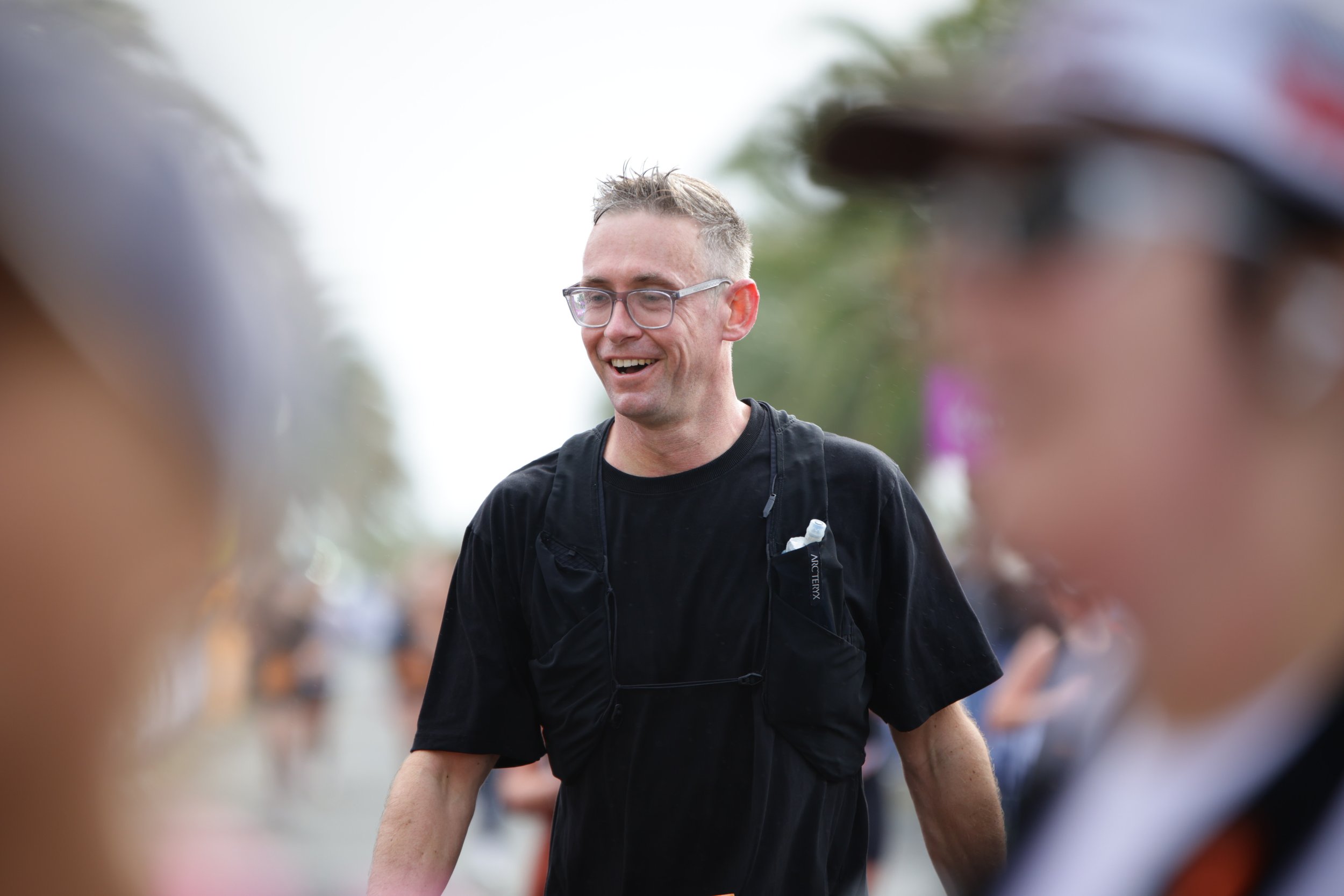 A man with glasses, short gray hair, and a black shirt smiling outdoors, surrounded by blurred figures of people.