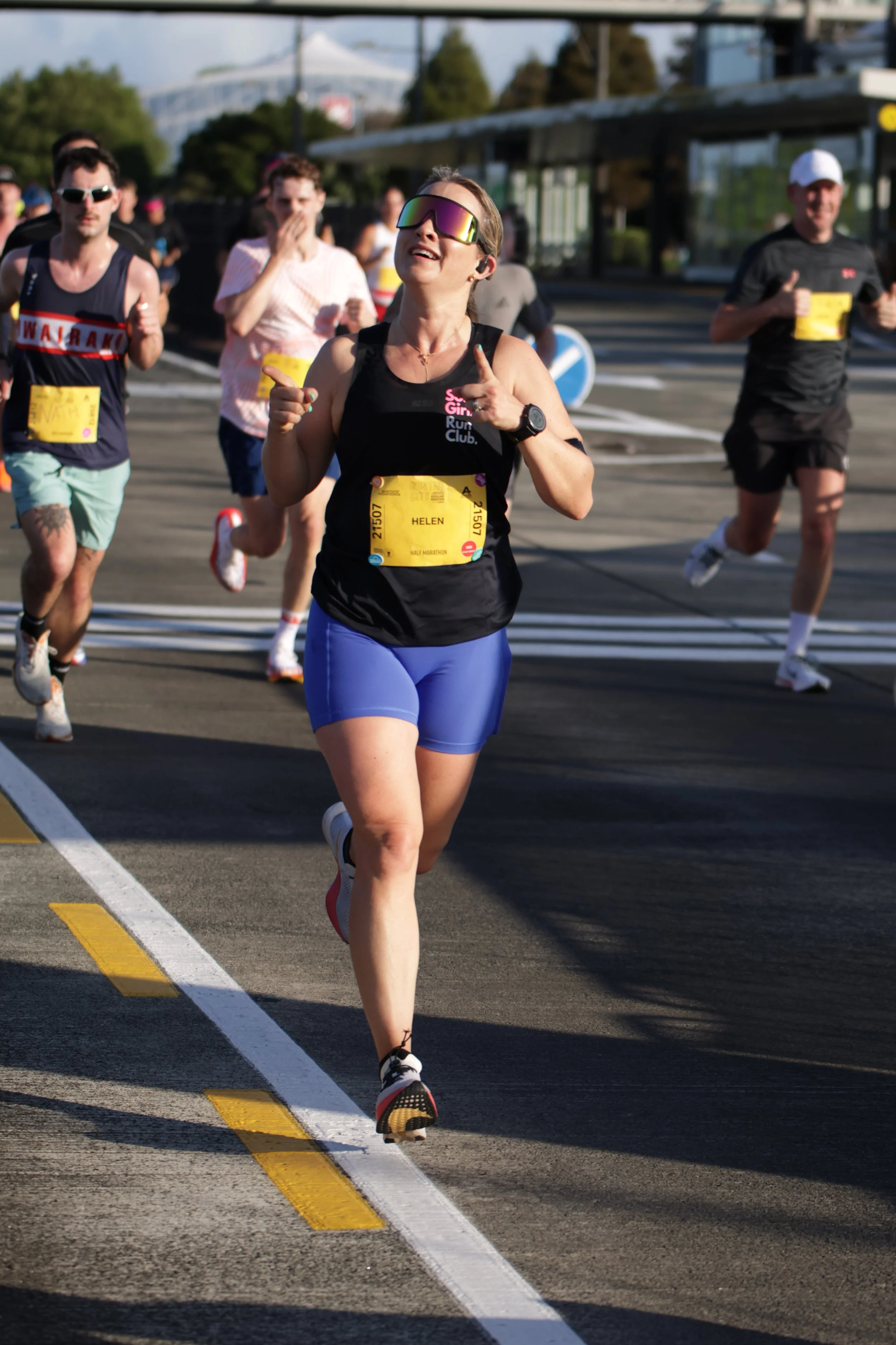 A group of runners participating in a marathon, with a woman in the foreground wearing sunglasses, a black sleeveless top, blue shorts, and a yellow bib number with the name Helen. The runners are on a city street with traffic signs and a tram stop i