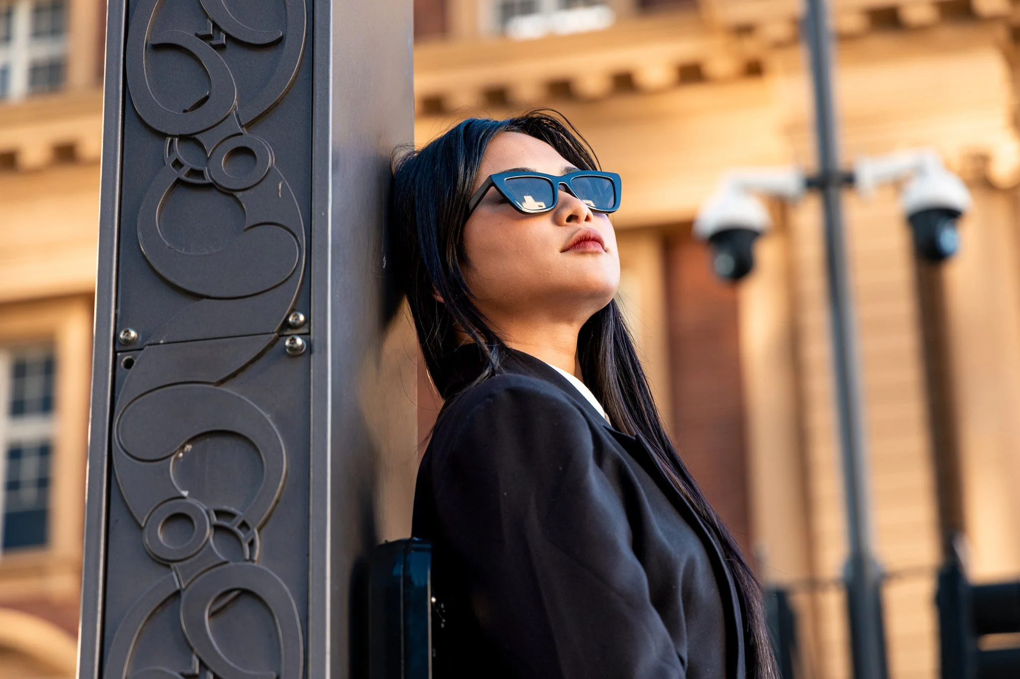 A woman with dark hair, wearing black sunglasses and a black jacket, leaning against a large metallic pole with circular designs, in an urban setting with a wooden building in the background.