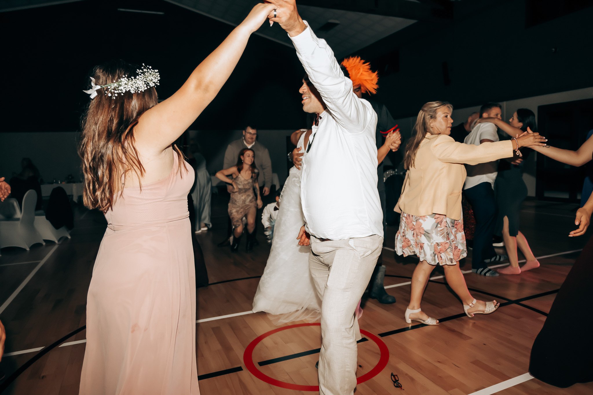 People dancing at a party in a gymnasium, with a woman in a pink dress and a woman in a beige skirt in the foreground, and other guests dancing in the background.