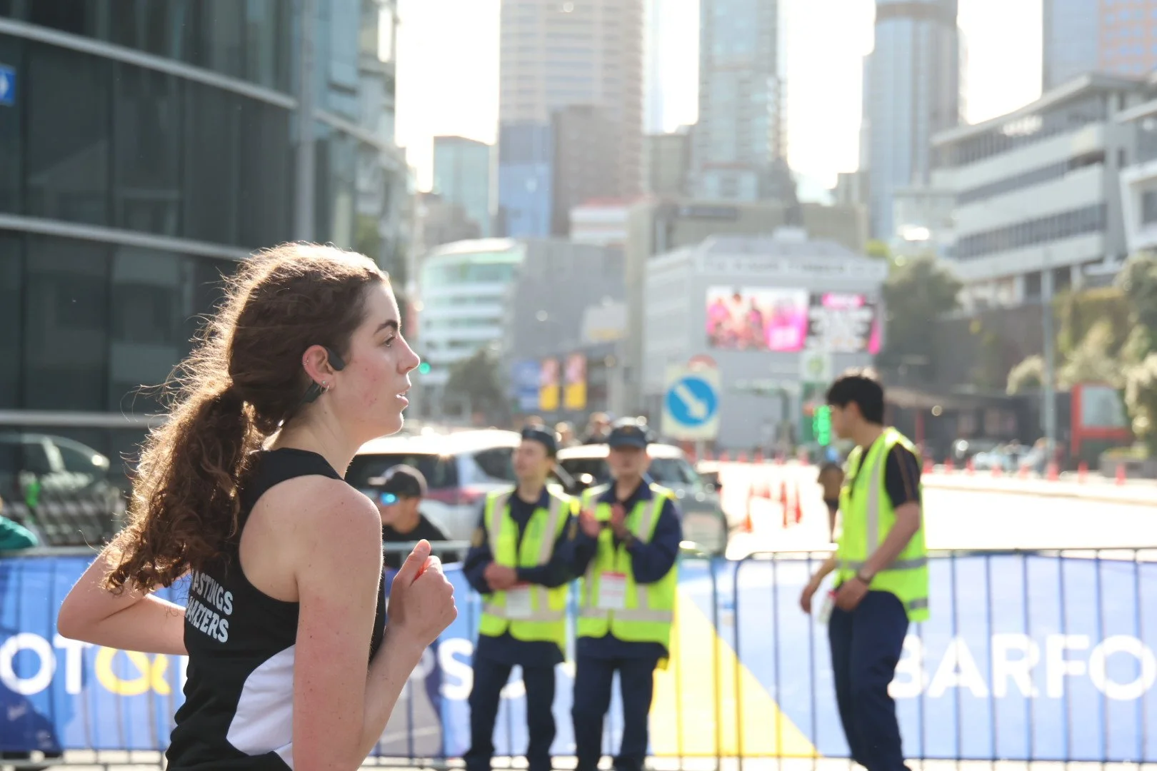 A female runner with curly hair and a black and white sports top runs in an urban setting as police officers in high-visibility vests stand in the background, with tall buildings and city streets behind them.