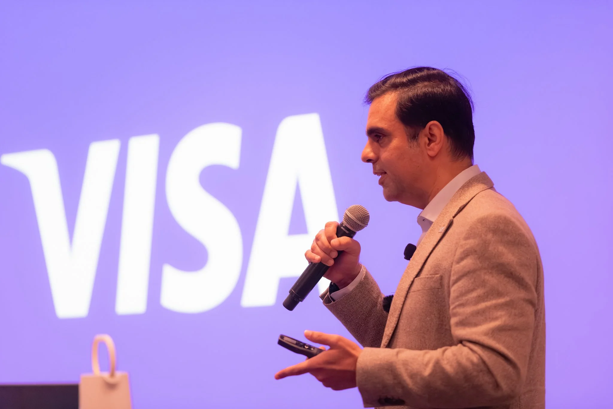 Man speaking into a microphone while holding a phone, in front of a purple background with the word "VISA" partially visible.