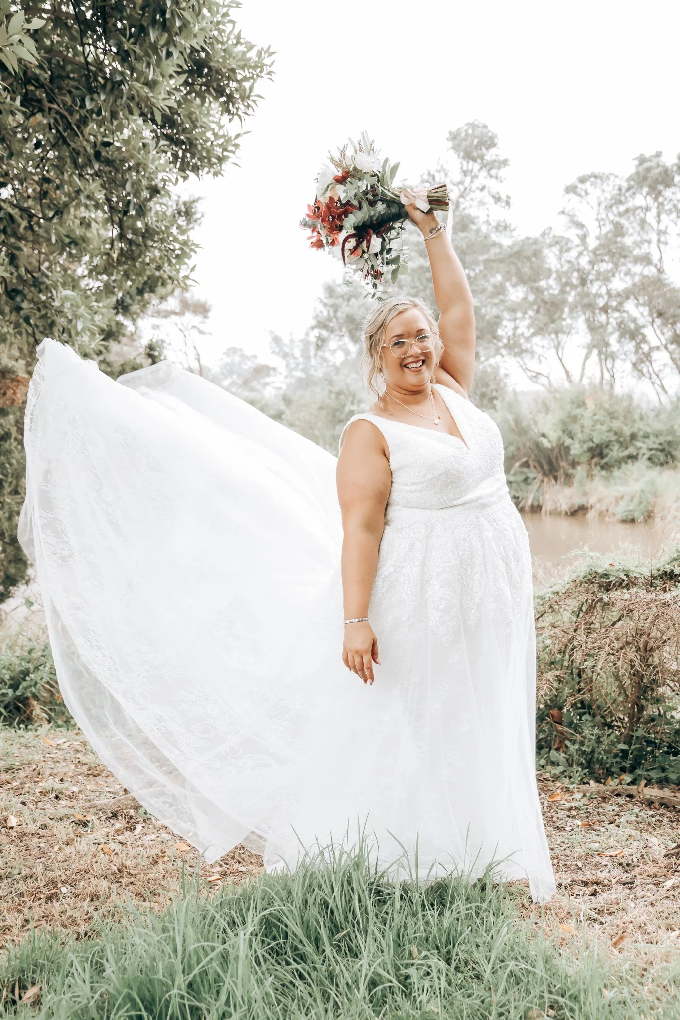 A woman in a wedding dress holding a bouquet of flowers above her head outdoors near a creek, with trees and grass in the background.