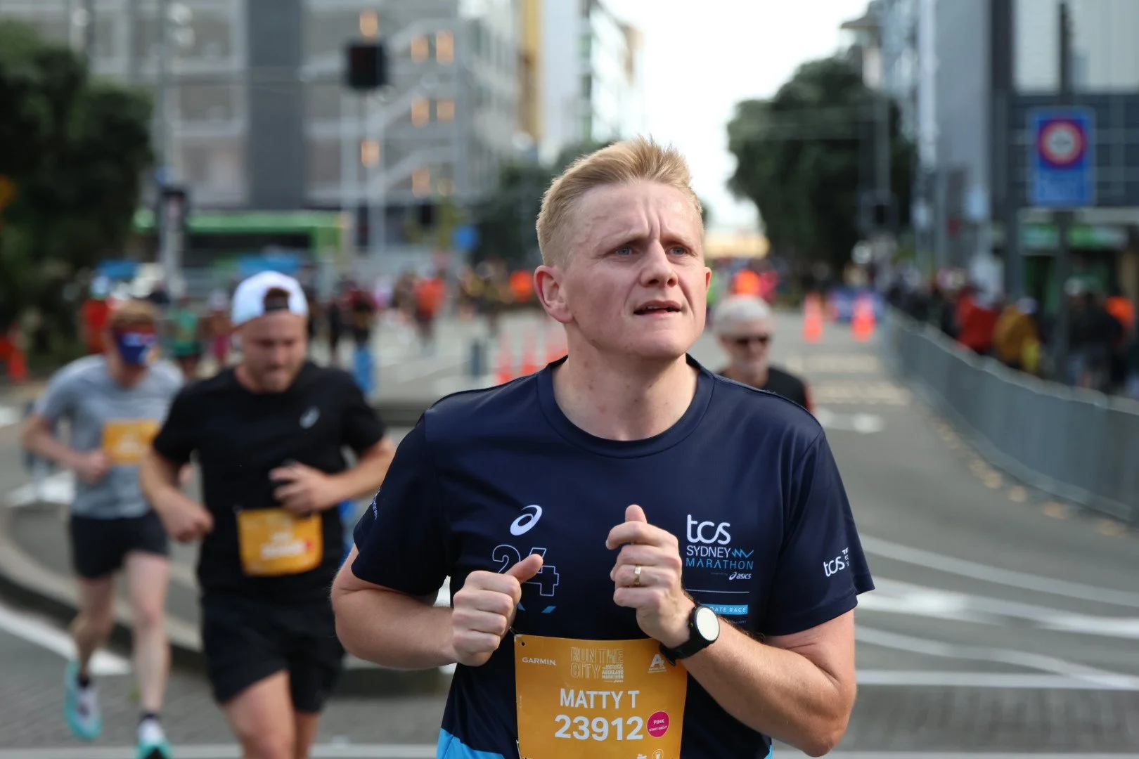 A man running in a marathon with a city street background, wearing a navy blue T-shirt with race branding and a bib labeled 'Matty T 23912'.