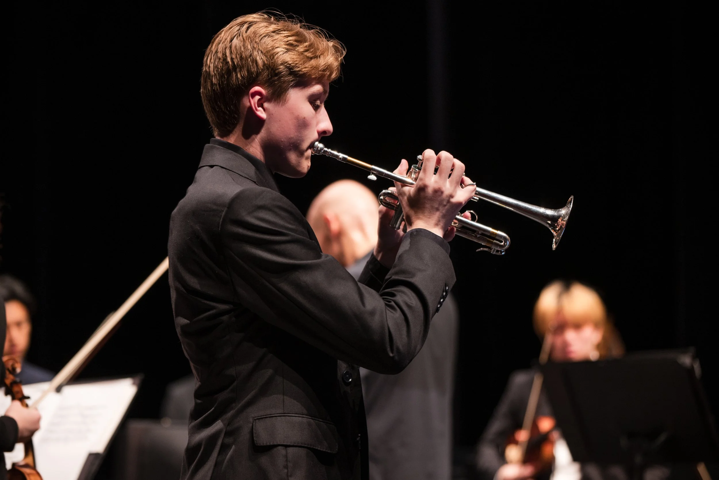 Young male musician playing the trumpet during a concert, wearing a black suit against a dark background.
