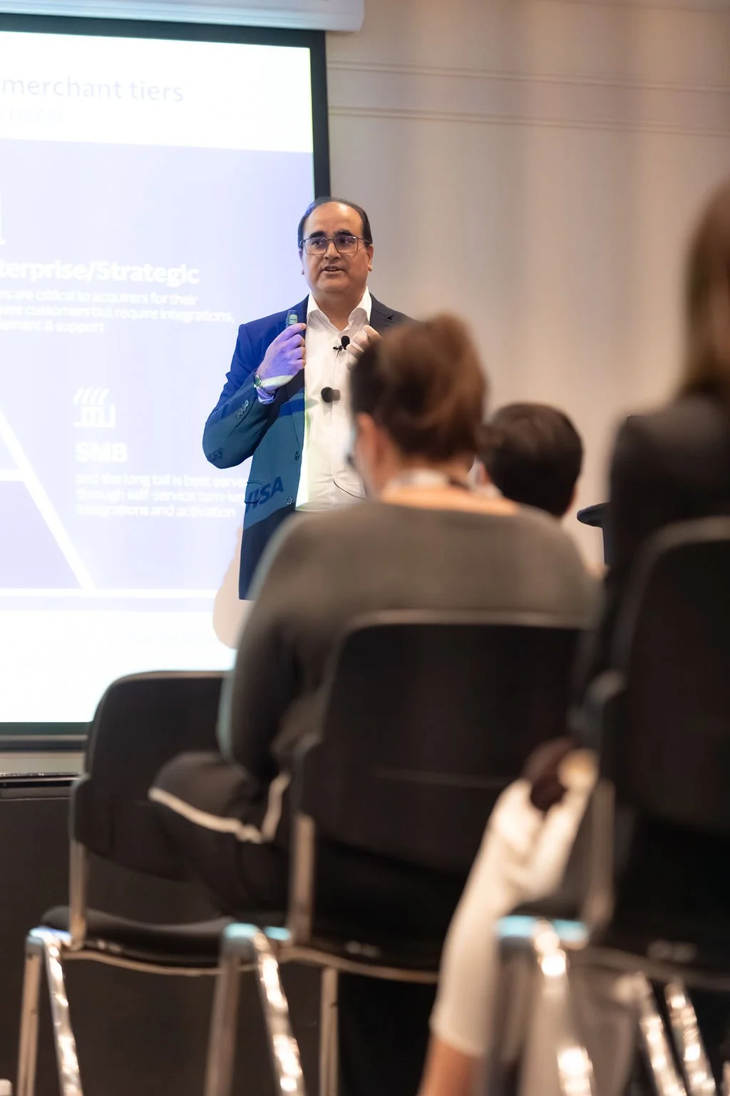 A man in a dark suit and glasses giving a presentation in front of a projected slide, with several audience members seated and listening.