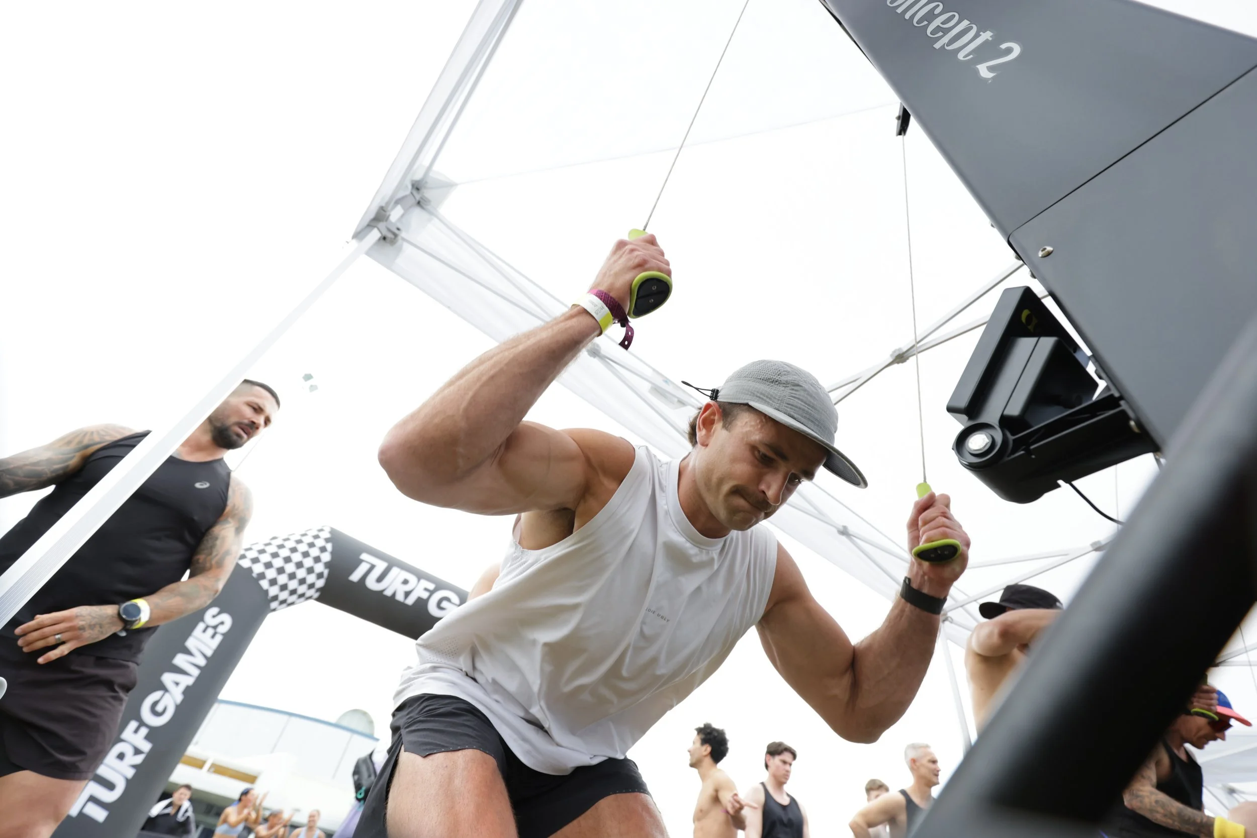 Two men preparing for a race under a canopy with a 'TURFG' banner, one holding a device, and a crowd of runners and spectators in the background.